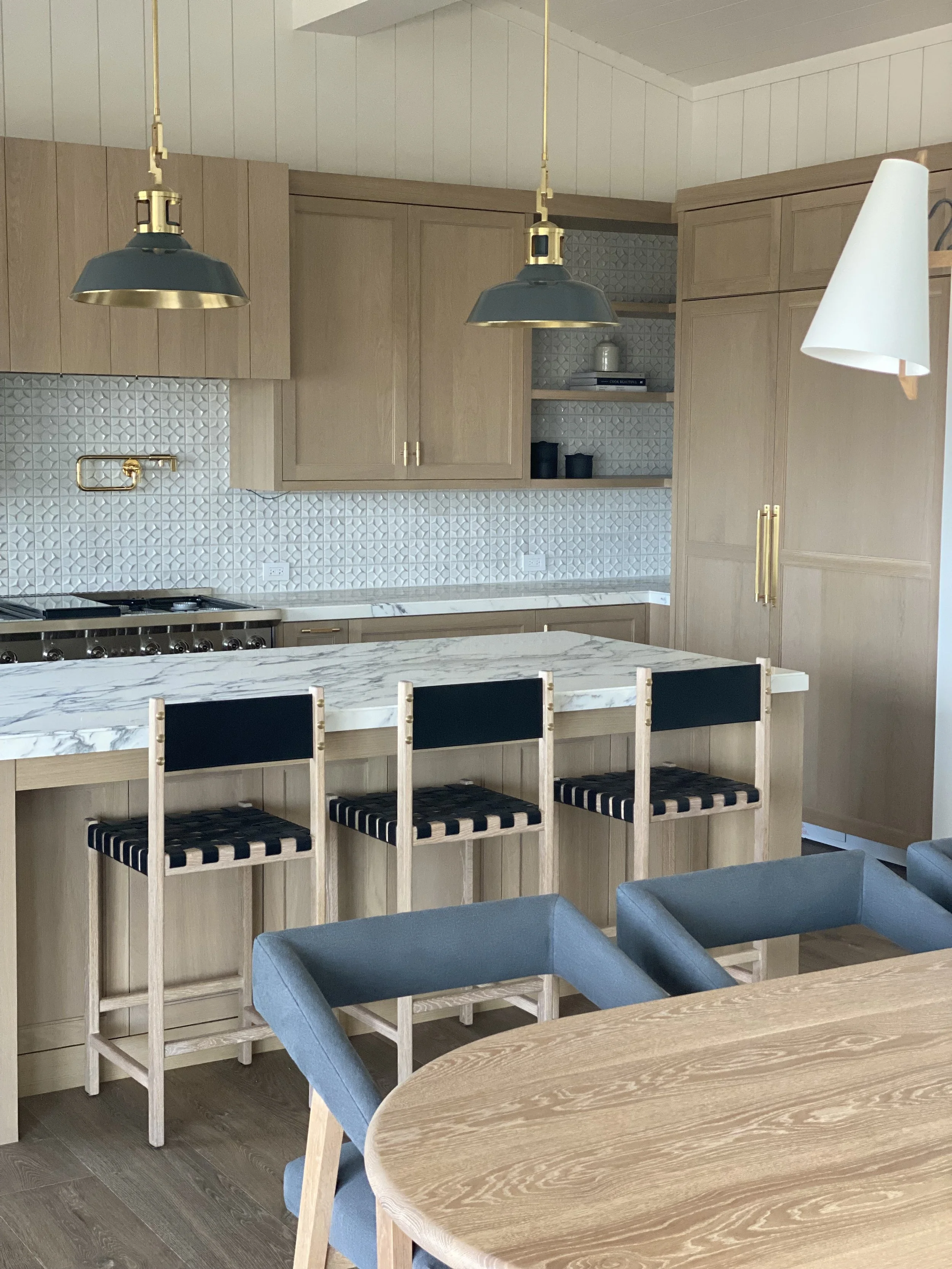 Modern kitchen with light wood cabinets, a marble countertop, blue and black bar stools, pendant lighting, and a dining table with blue chairs.