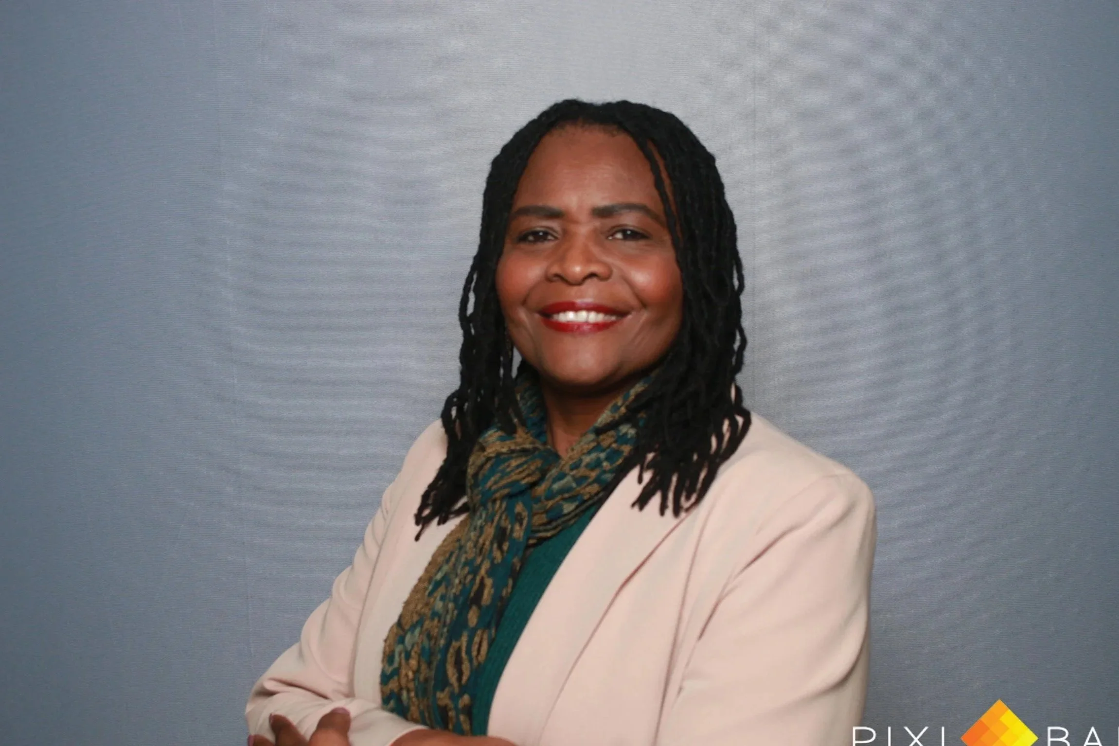Portrait of a smiling woman with black hair styled in dreadlocks, wearing a beige blazer and a colorful scarf, against a light blue background.