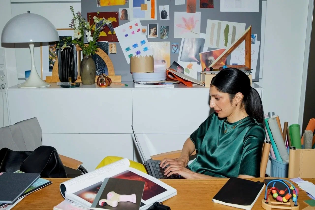 Woman working on a laptop at a cluttered desk with art supplies and decor, in a room with a white cabinet and a bulletin board covered with art and photos.