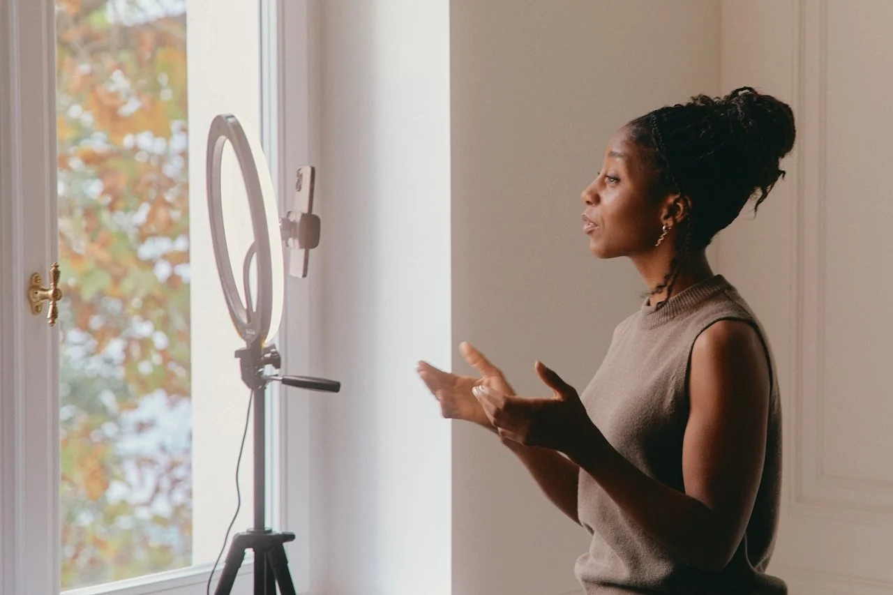 Woman using a ring light for a video call or recording in a room with a window showing autumn leaves outside.