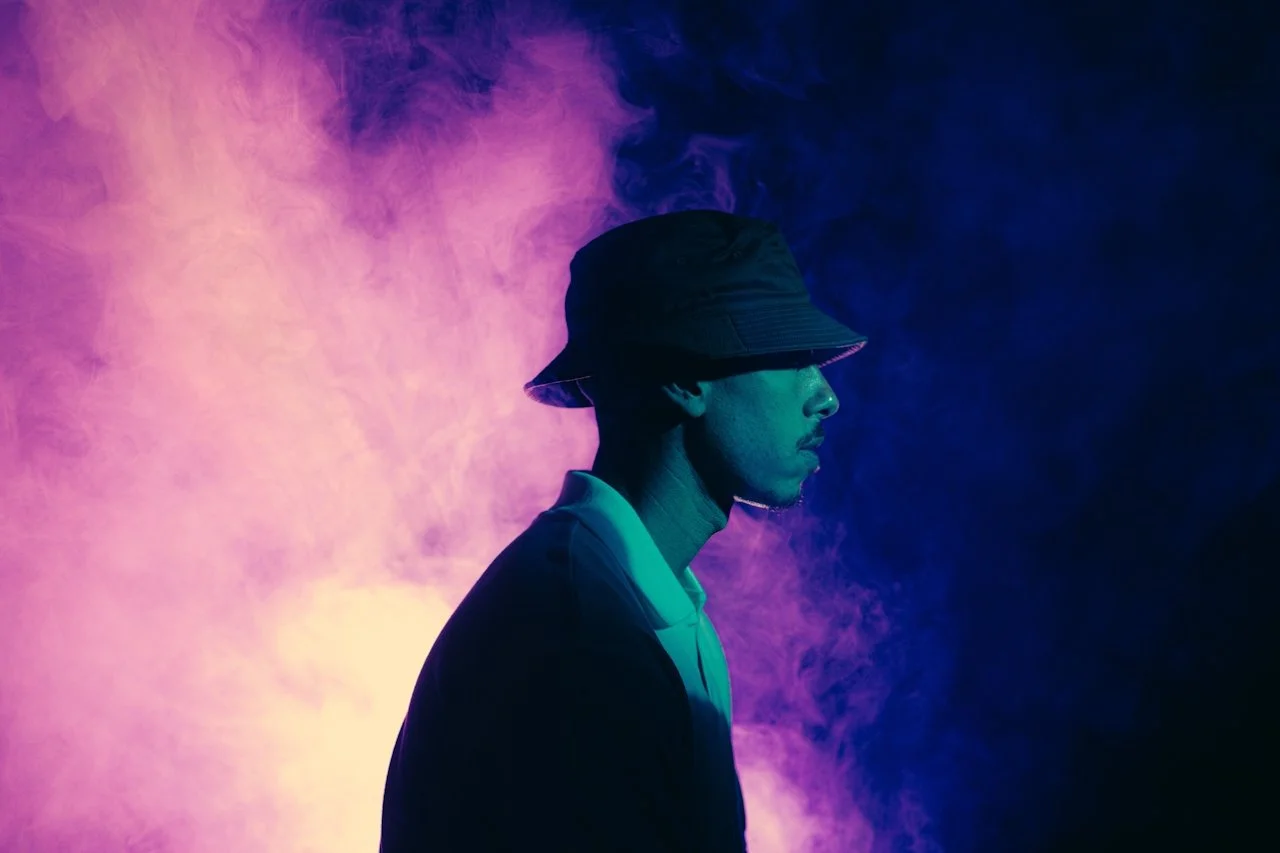 Side profile of a person in a bucket hat and jacket, standing in front of colorful purple and blue smoke.