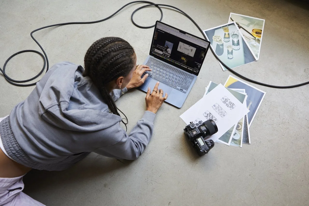 A woman lying on the floor working on a laptop surrounded by printed photographs and a camera.