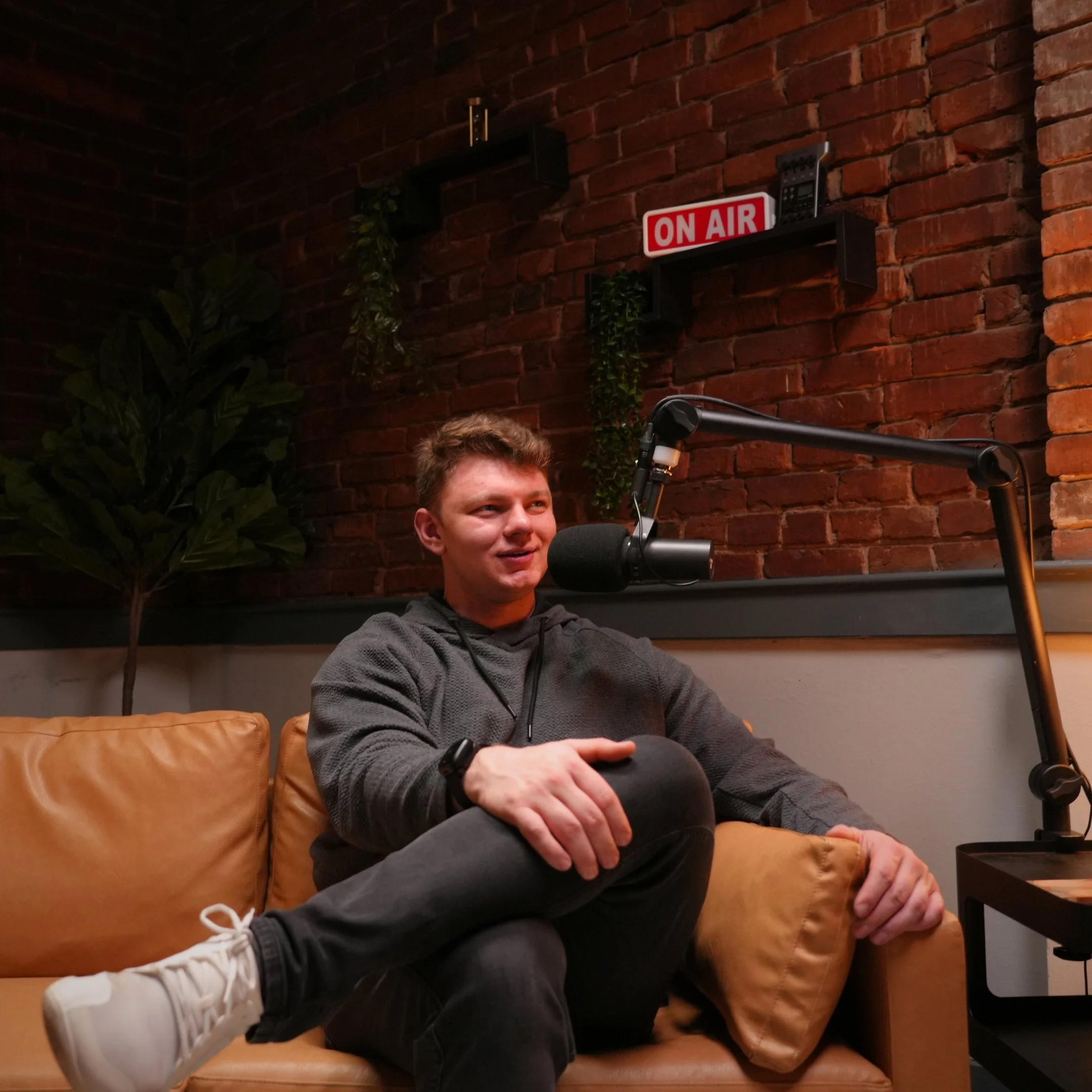 A young man sits on a tan leather couch in a recording studio or radio station, speaking into a microphone. Behind him, a brick wall features an illuminated 'On Air' sign, along with some decorative plants and broadcast equipment.