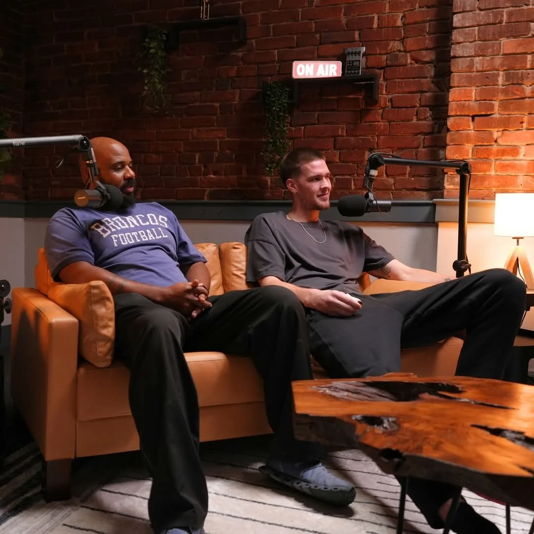 Two men sitting on a tan leather couch in a radio or podcast studio with brick wall background. One man wears a Broncos football t-shirt, and the other wears a gray t-shirt. A microphone is positioned in front of the man on the right. An 'On Air' sig