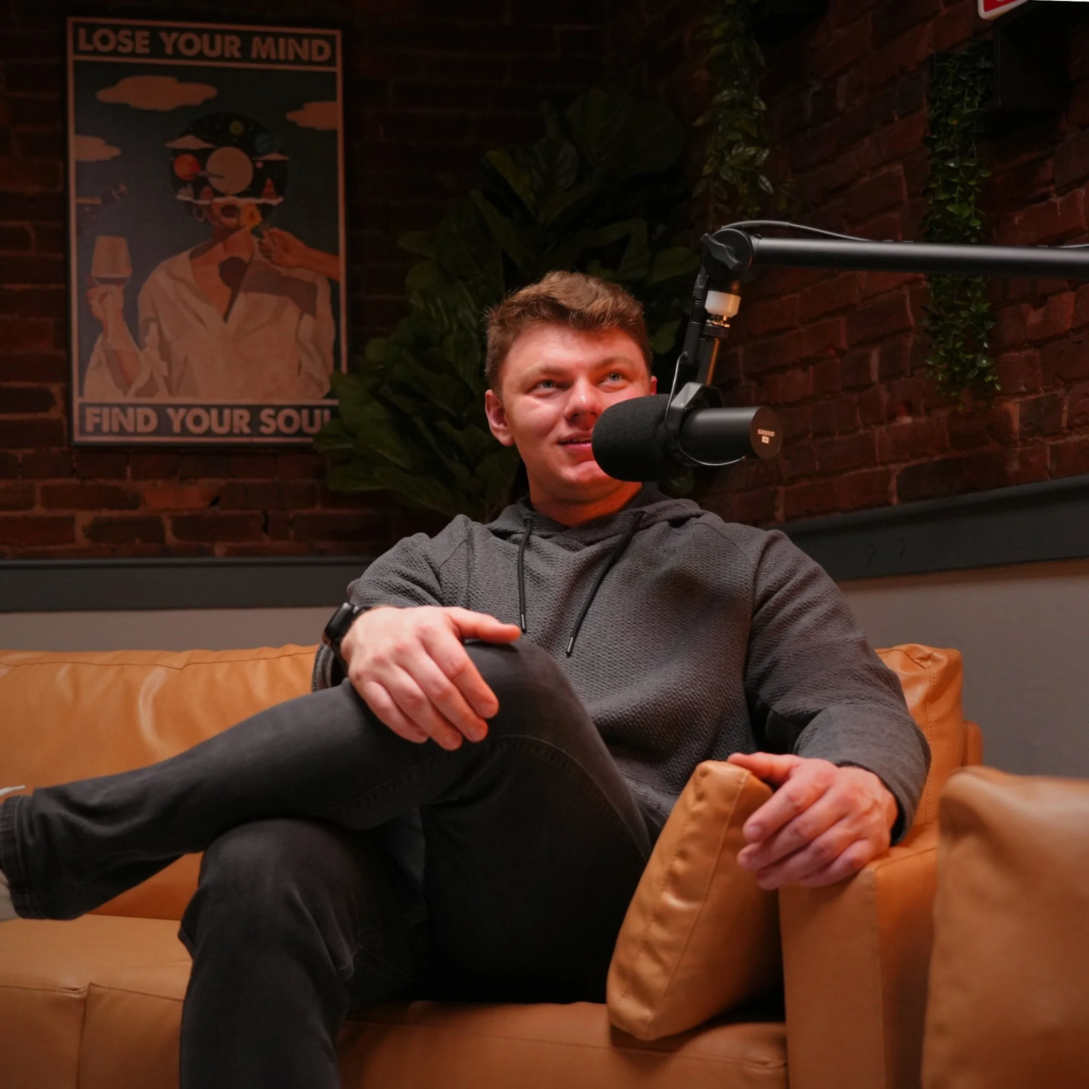 A young man sitting on a tan couch, speaking into a microphone, wearing a gray hoodie, with a brick wall and framed artwork in the background.