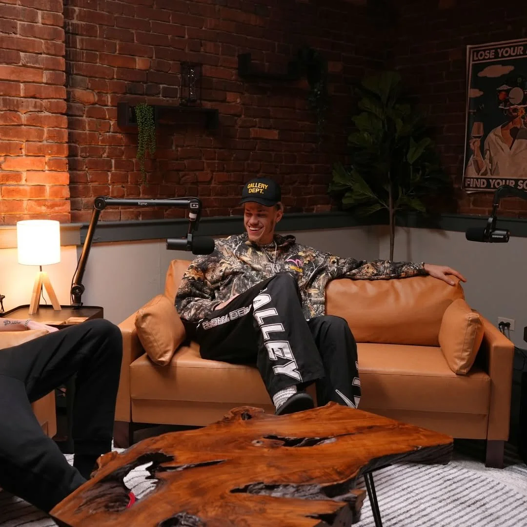 A young man sitting on a tan leather sofa, wearing a camouflage jacket, black pants with white lettering, and a black cap that reads 'GALLERY DEPT.' He is smiling and appears to be having a conversation in a podcast studio. The studio has a brick wal