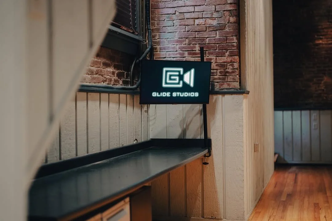 Neon sign on wall reading 'GLIDE STUDIOS' with a camera icon, inside a room with brick and wood walls and hardwood floor.