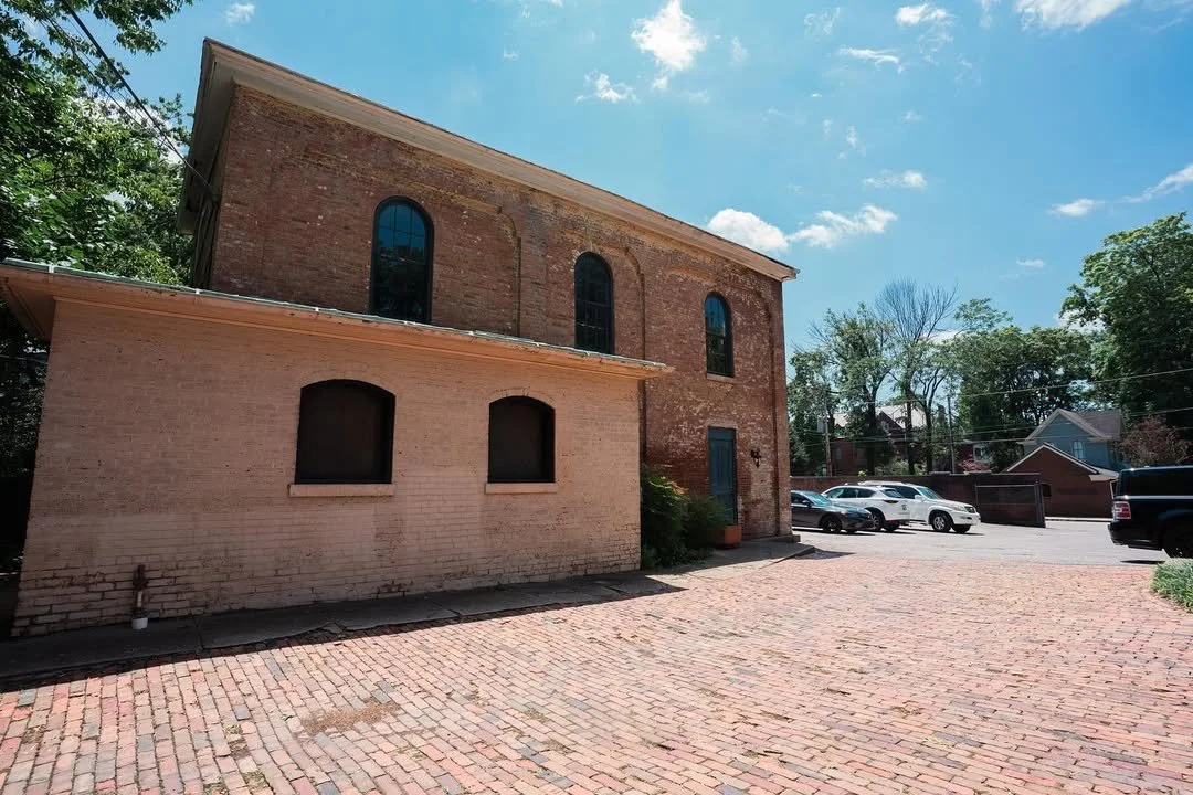Brick building with arched windows, a small brick extension, a brick patio, and parked cars under a partly cloudy blue sky.