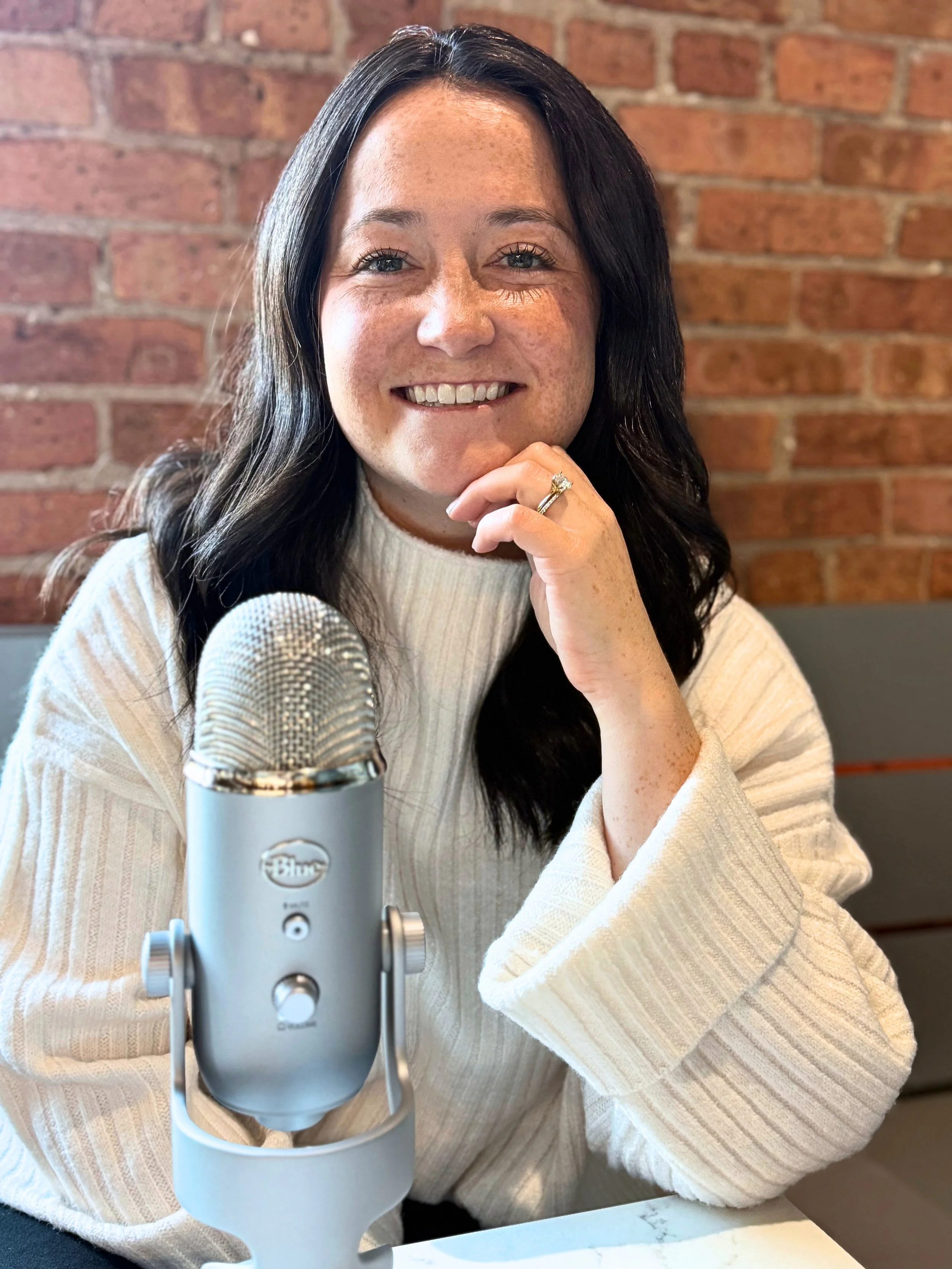 A woman with long dark hair, wearing a white ribbed turtleneck sweater, sitting at a table with a silver Blue Yeti microphone in front of her, smiling and resting her chin on her hand, in front of a brick wall.