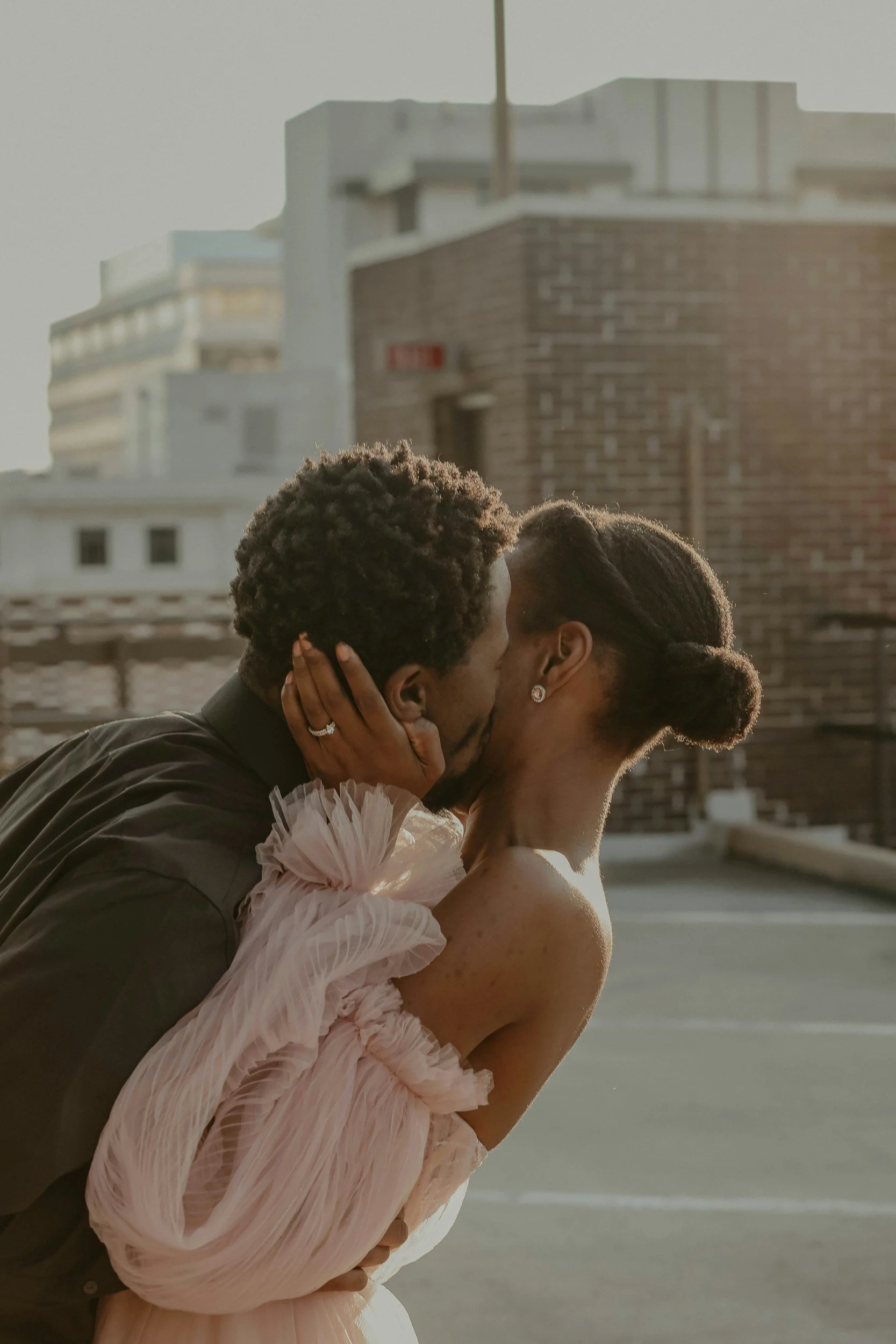 A couple sharing a kiss on a rooftop in the late afternoon, with city buildings in the background.