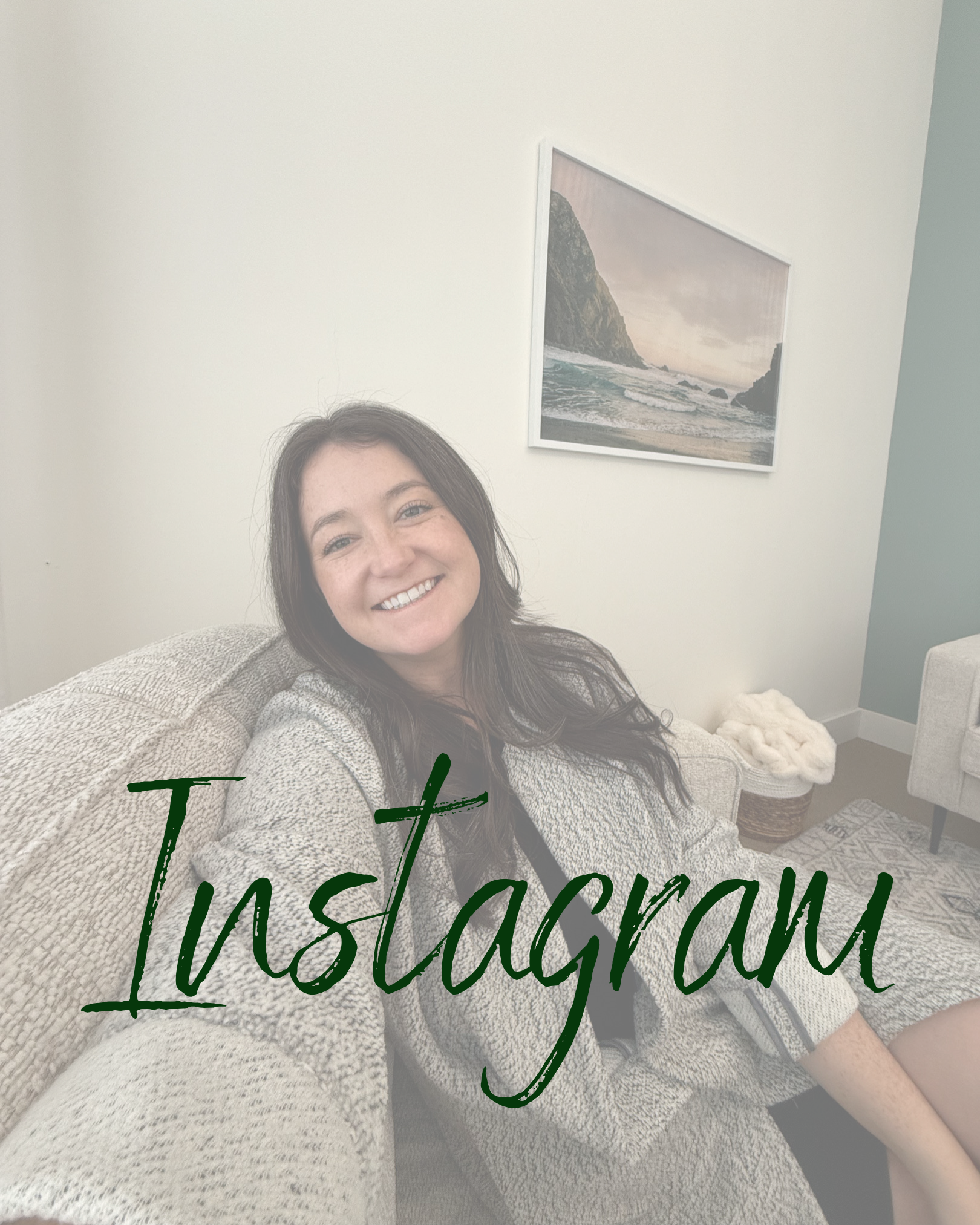 A woman with long brown hair smiling while sitting on a beige couch in a living room with light-colored walls, a framed picture of a mountain and ocean scene, and a basket with a fluffy blanket.