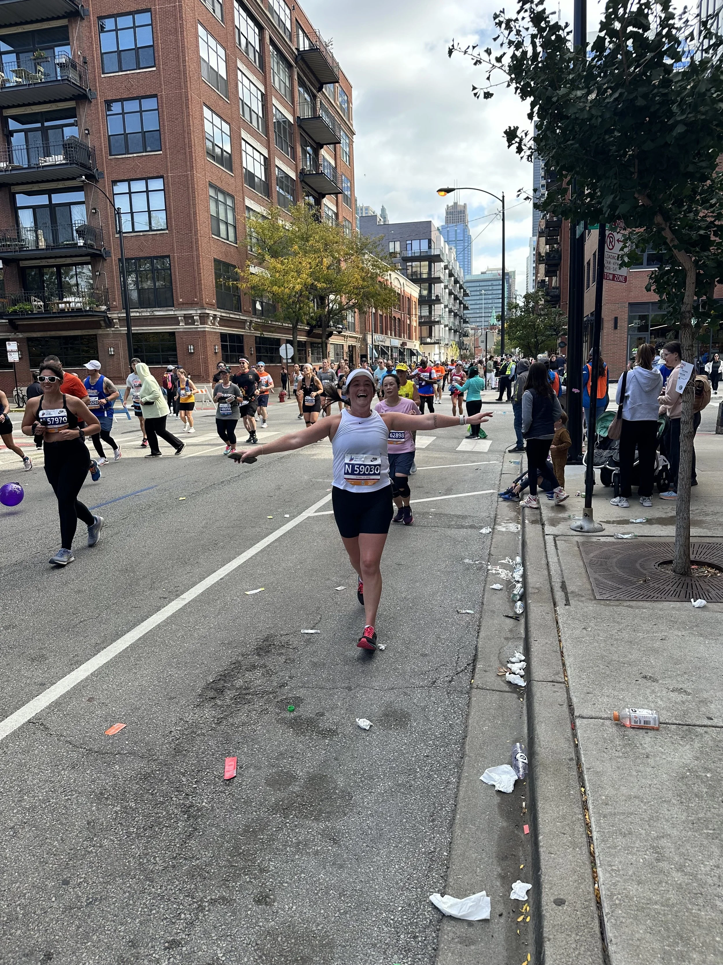 A joyful woman running in a marathon on a city street, surrounded by other runners and spectators, with tall buildings and trees in the background.