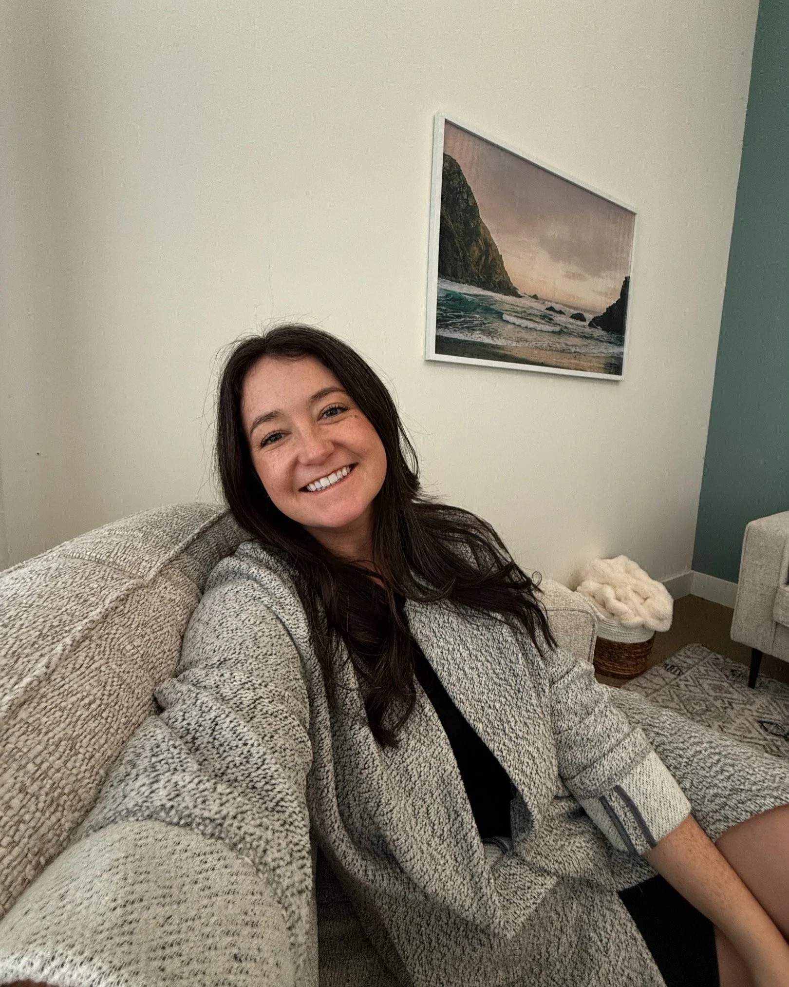 A woman with long dark hair smiling and sitting on a beige sofa in a living room