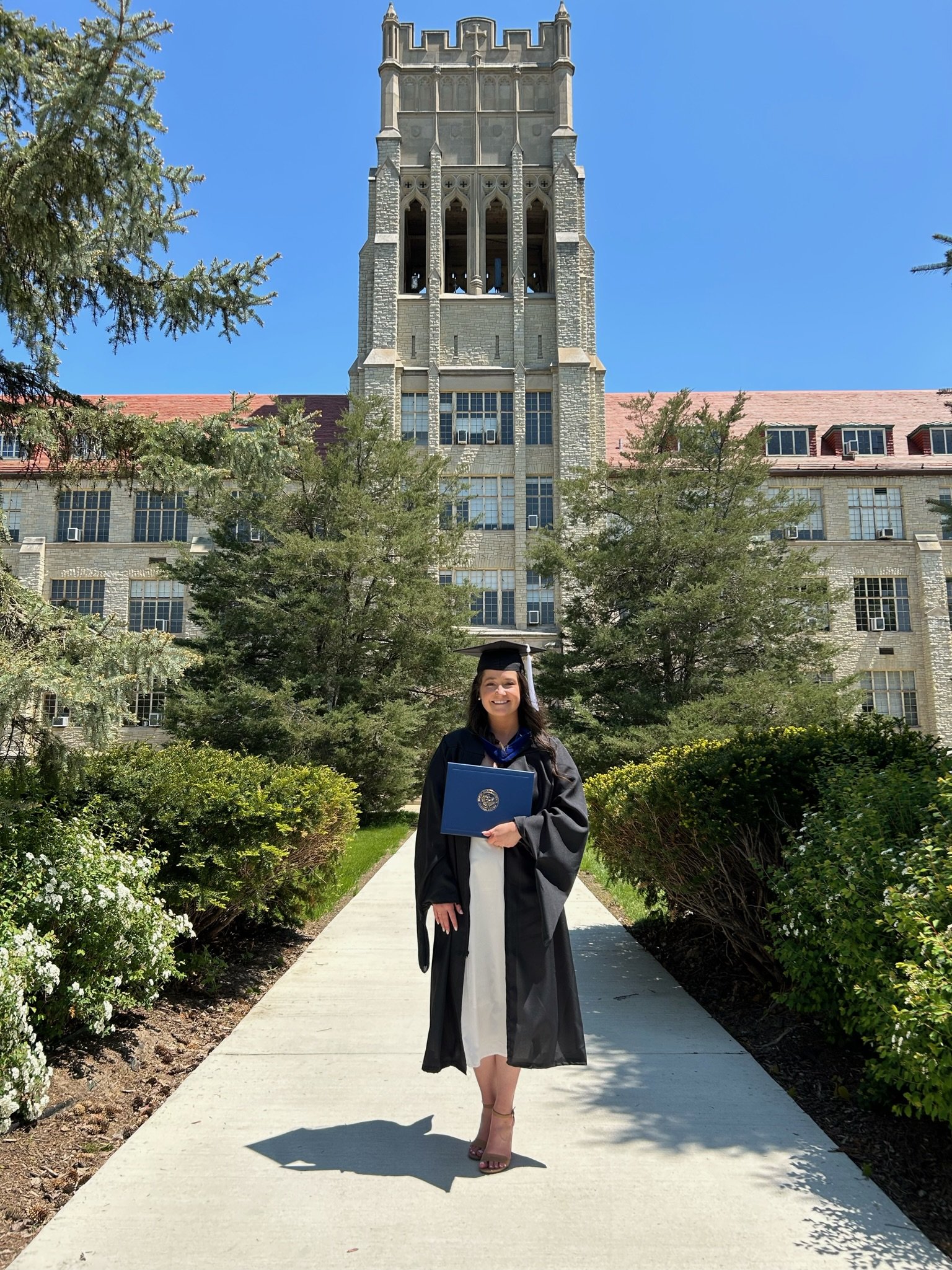 A graduate in cap and gown holding a diploma, standing on a walkway in front of a large university building with a tower, surrounded by trees and bushes, on a sunny day.