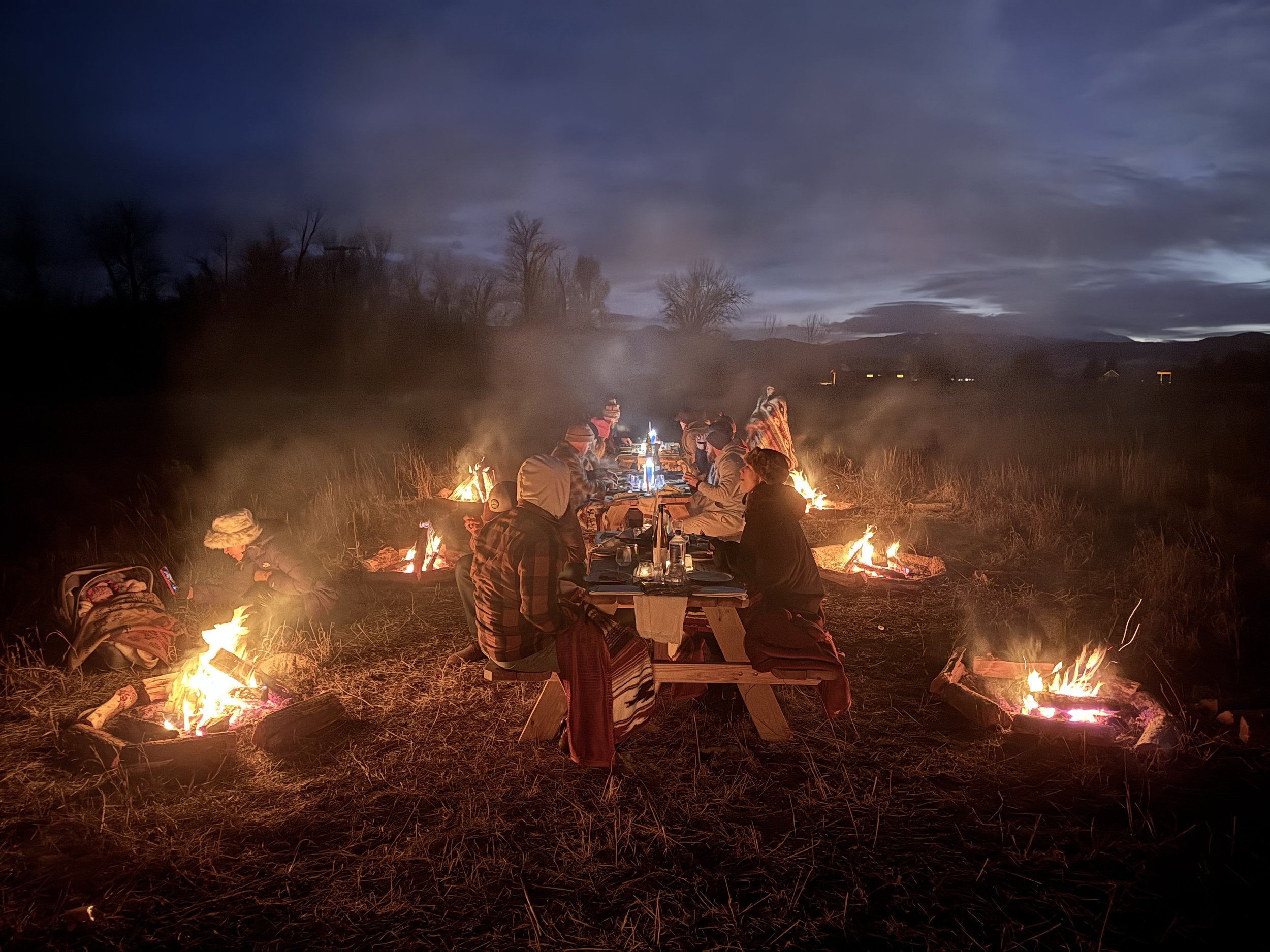 Group of people sitting at a long picnic table outdoors at night, surrounded by small campfires, with a cloudy sky and mountains in the background.