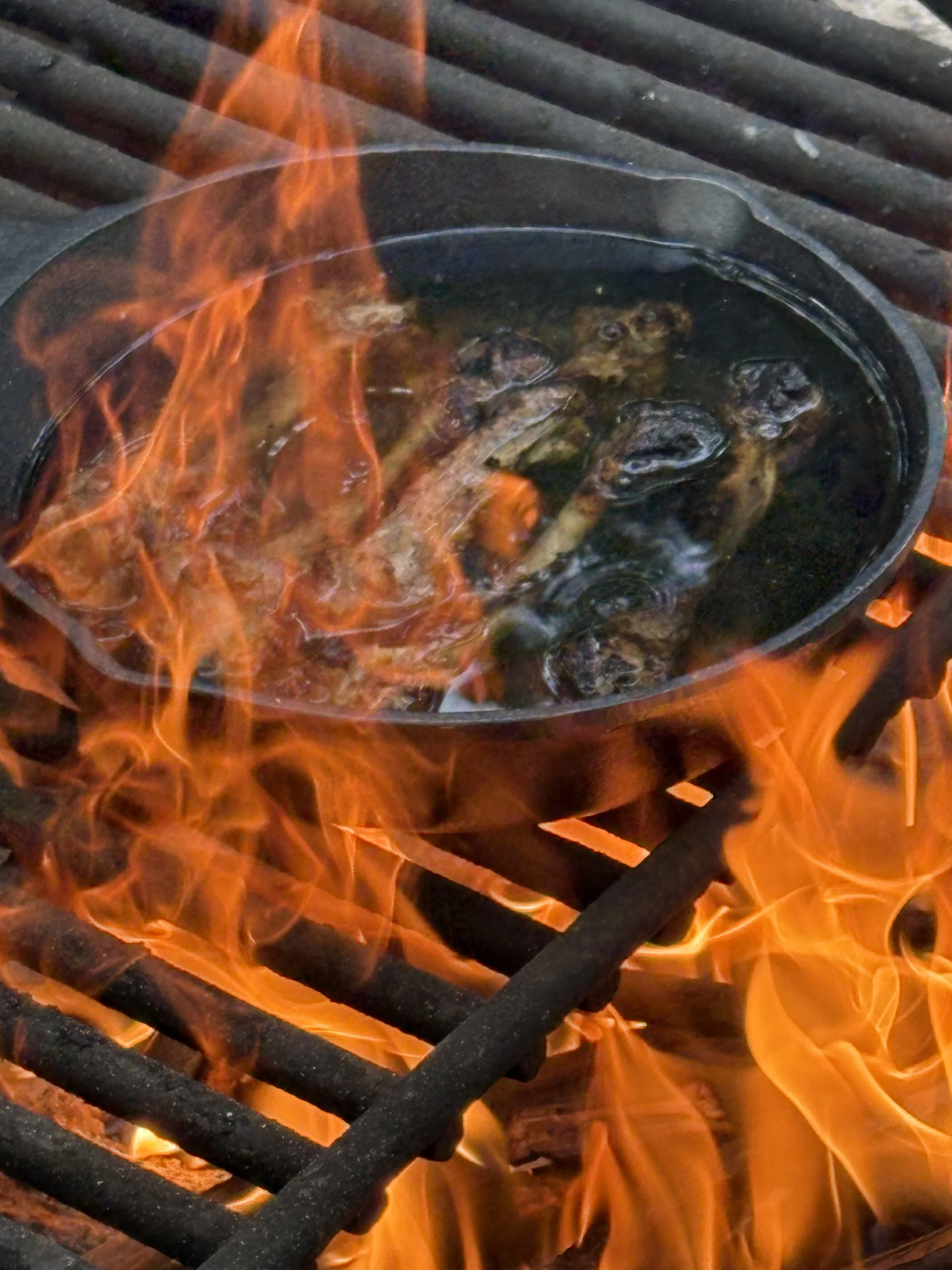 A cast iron skillet with food inside, placed on a grill over an open flame with orange and yellow fire.