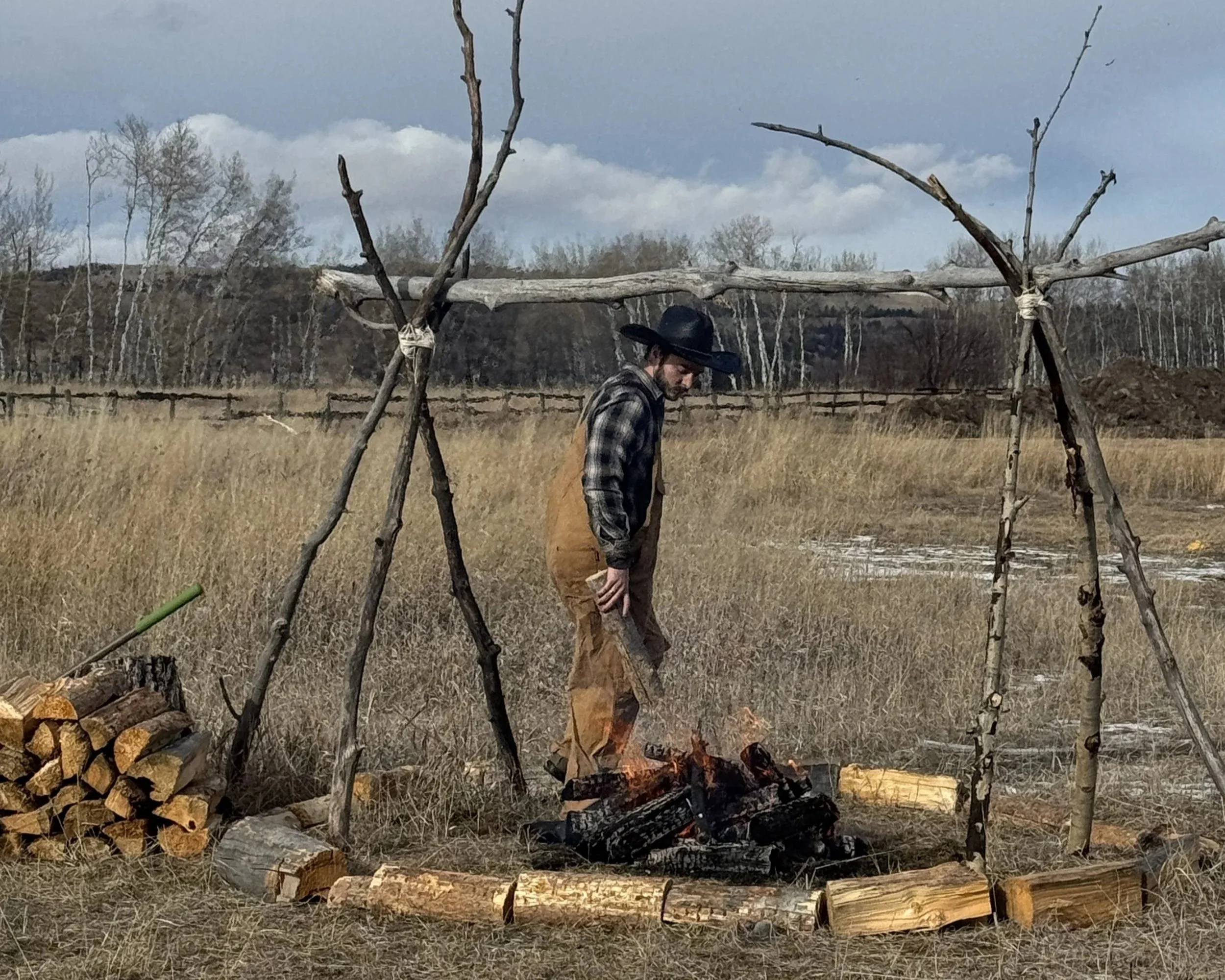A man in plaid shirt, brown overalls, and black hat tending a fire inside a makeshift teepee made of tree branches in a grassy field with stacks of wood nearby.