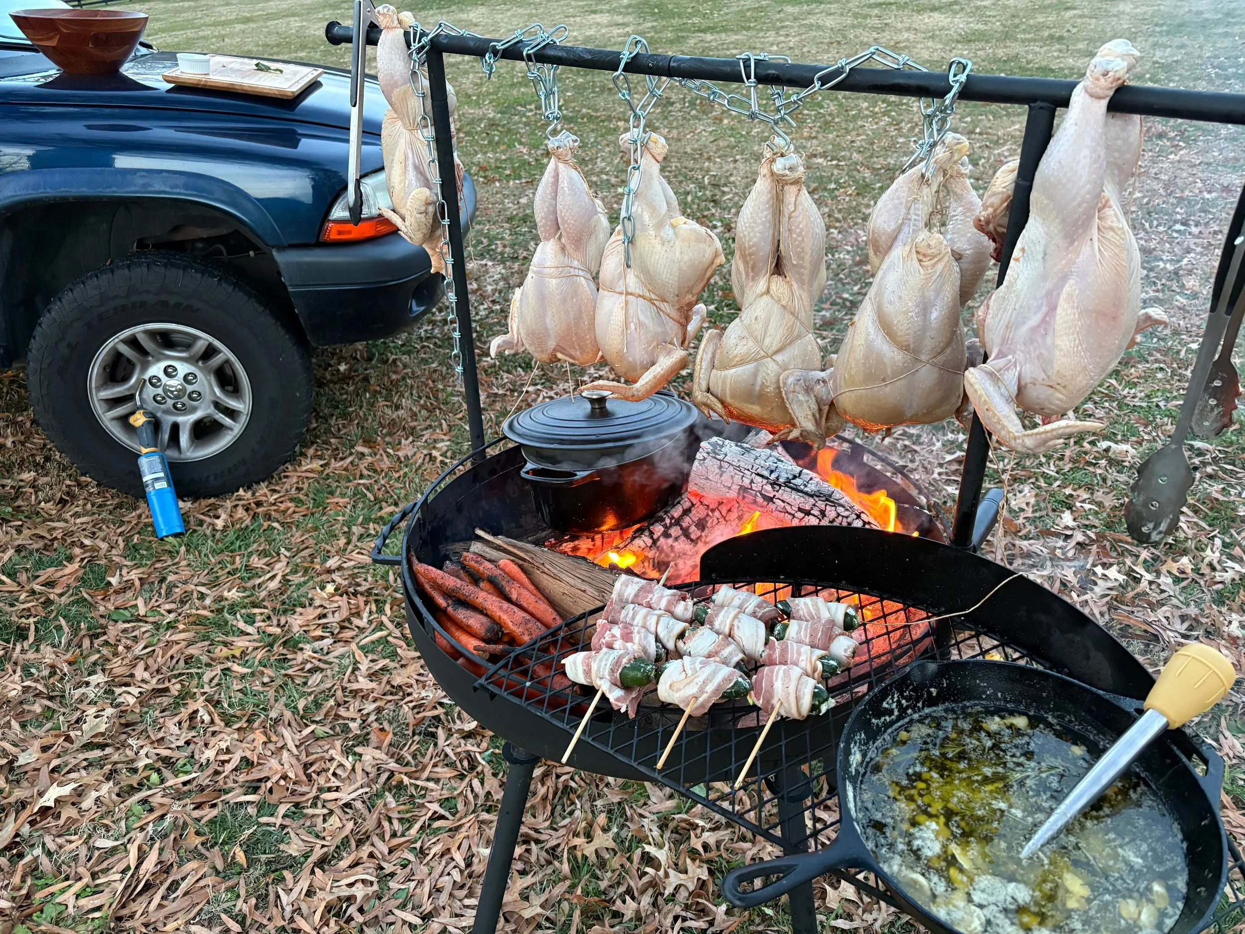 Barbecue grill with whole chickens hanging on a rack, skewered bacon-wrapped jalapenos on skewers, and hot dogs cooking on the grill. A pan with melted butter and herbs is on the grill, with an outdoor wooded area and a parked blue SUV in the background.