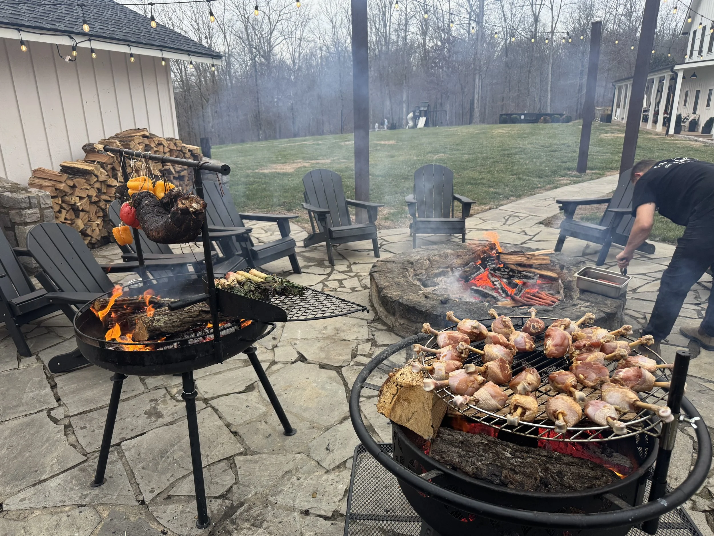 Outdoor backyard scene with three portable grills on a stone patio, one with skewered chicken, another with firewood and hanging vegetables, and a person tending to the grill. There are several black Adirondack chairs, a woodpile against a building, and string lights overhead. In the background, there is a grassy area with trees and a house.