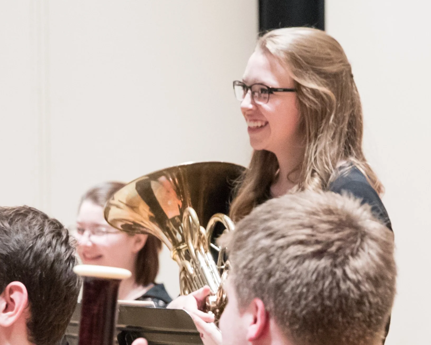 A woman with glasses smiling while holding a shiny brass musical instrument, likely a French horn, in a group setting with other people.