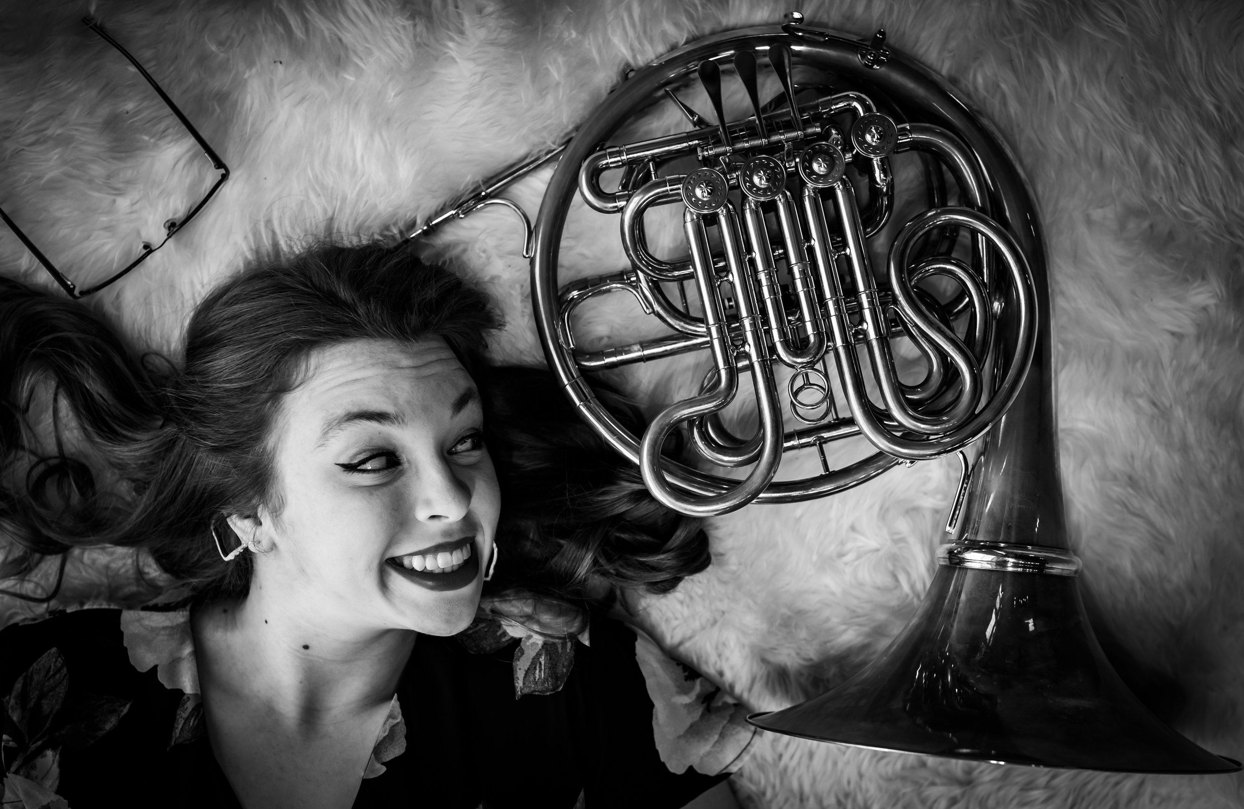 A woman lying on a fluffy surface, smiling and winking at the camera, with a French horn and a pair of glasses nearby.