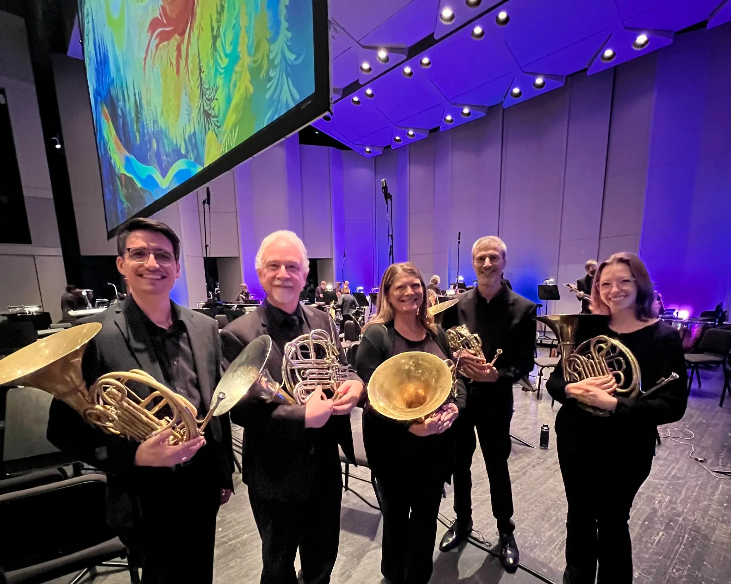 Five musicians with French horn brass instruments, standing on stage in a concert hall with a large screen displaying firebird abstract art behind them.