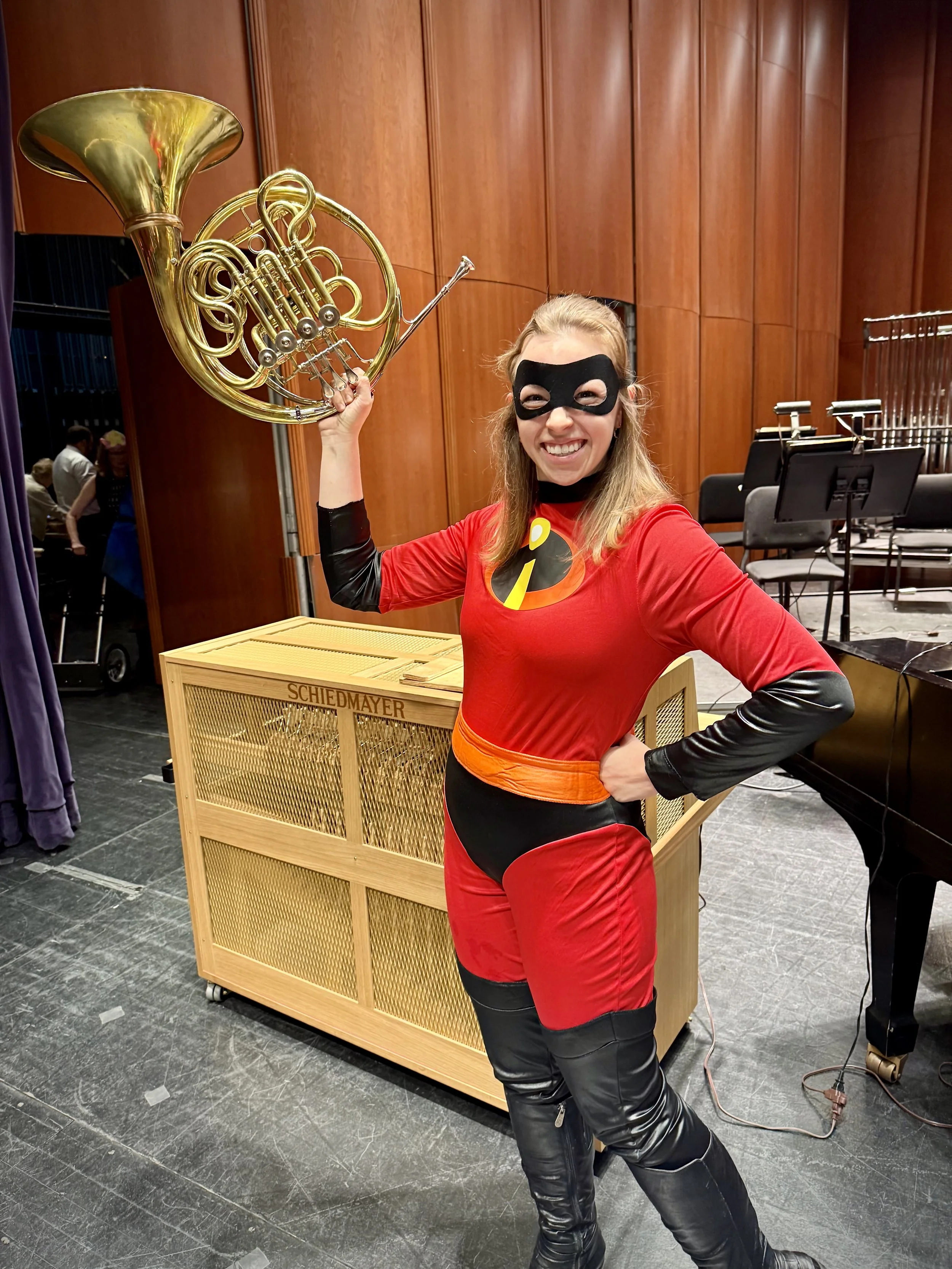 A woman dressed as Elastigirl from The Incredibles, wearing a red and black superhero costume with an elastigirl logo on her chest, black eye mask, and black thigh-high boots, holding a shiny golden French horn in a music hall or stage setting.
