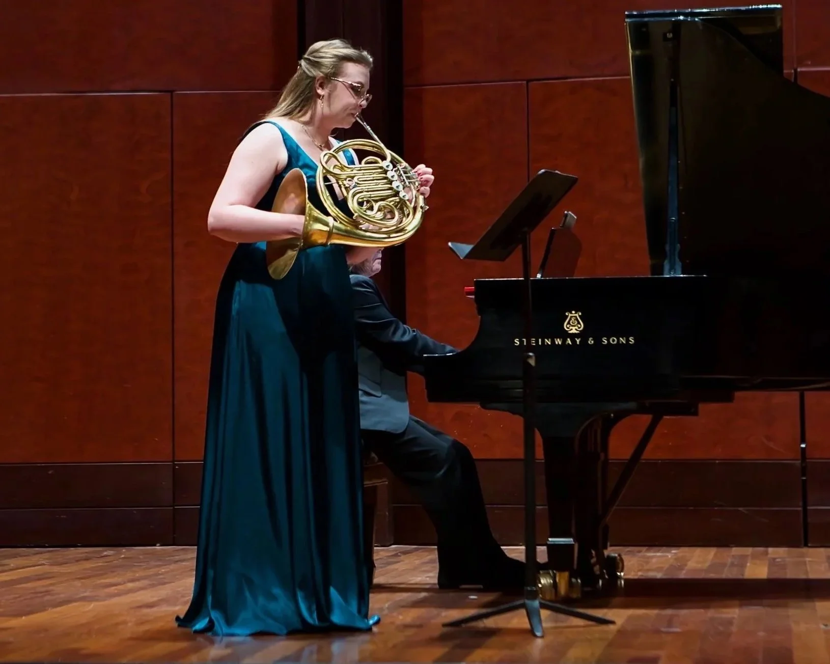 A woman in a long green dress playing the French horn while standing next to a woman playing the piano on a wooden stage with a red-brown wall background.