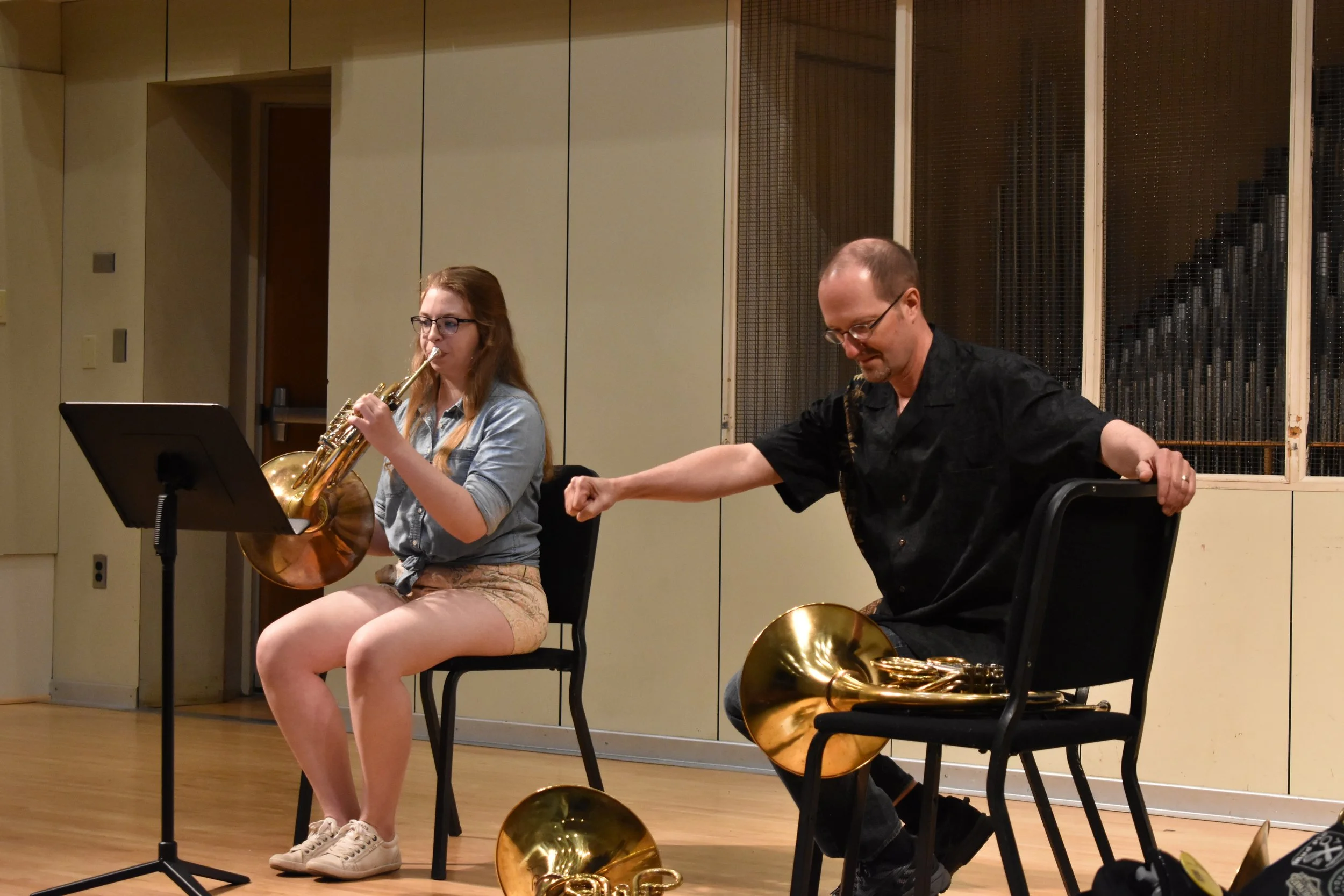 A young woman playing a French horn and a man sitting on a chair with a French horn in a music practice room recital hall