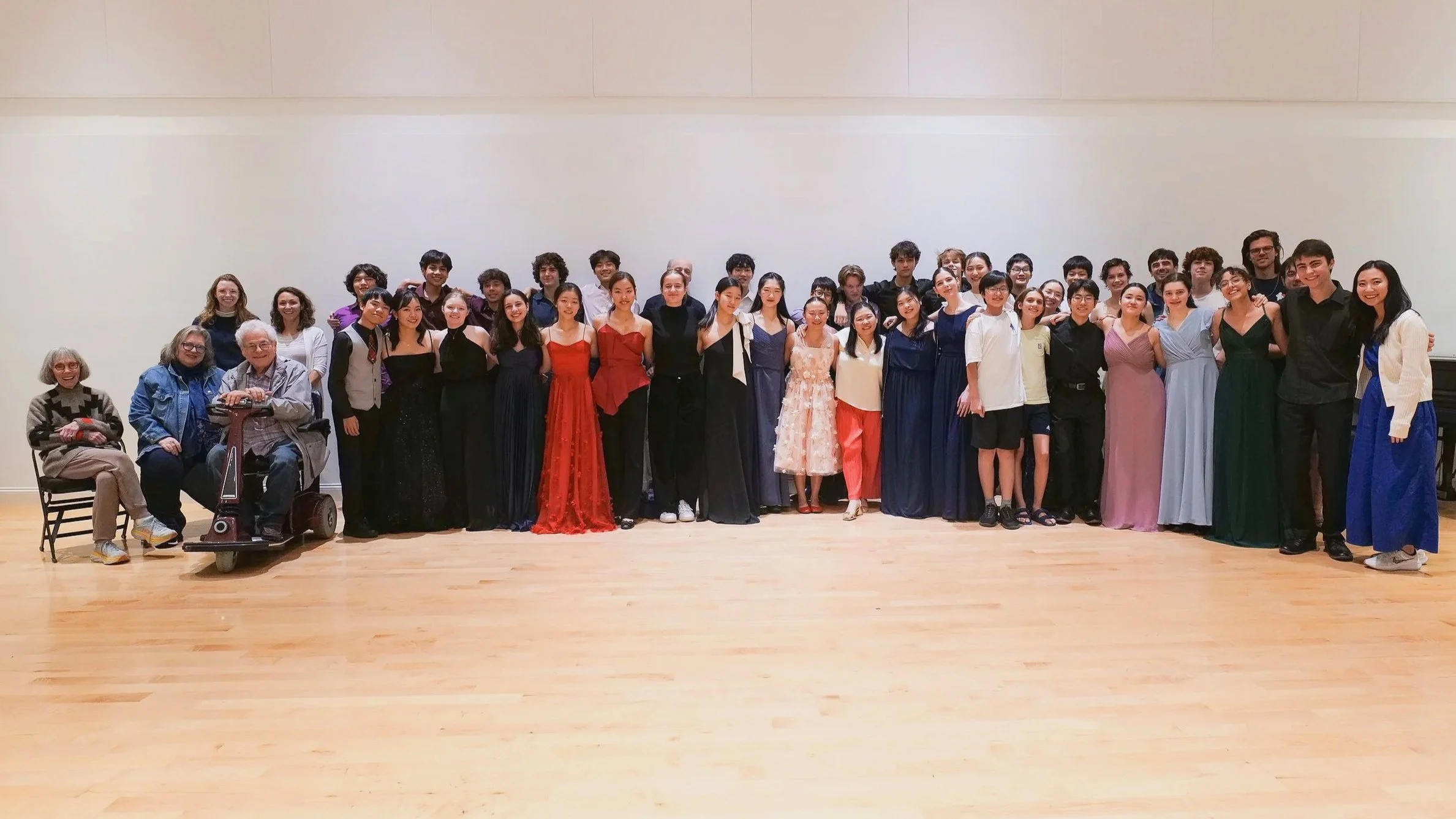 Large group of people, including musicians and performers, pose together in a spacious hall for a group photo. Some are dressed in formal evening gowns and tuxedos, while others are in casual attire. There are three individuals seated on the left side, including elderly people, one in a motorized wheelchair. The background is a plain white wall, and the floor is wooden.