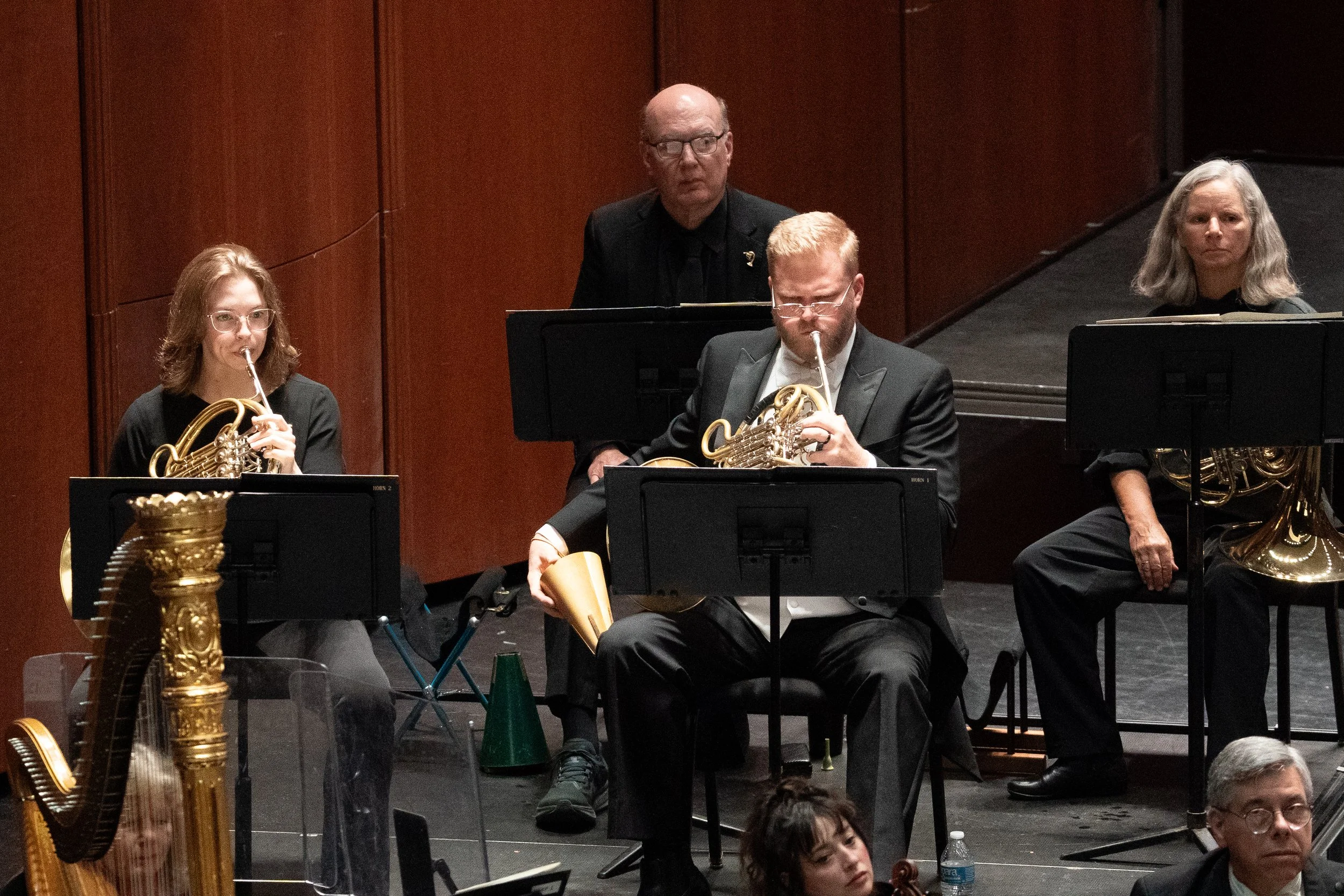 A group of musicians, including two women and two men, playing French horn musical instruments during a performance on stage with a brown wooden background.