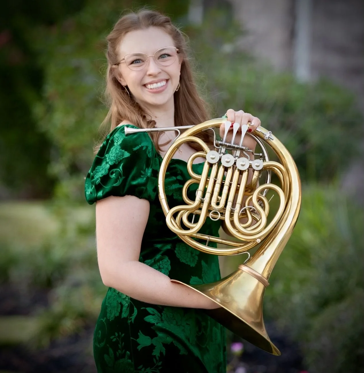 A young woman with red hair, glasses, and a green floral dress holding a French horn outdoors, smiling at the camera.