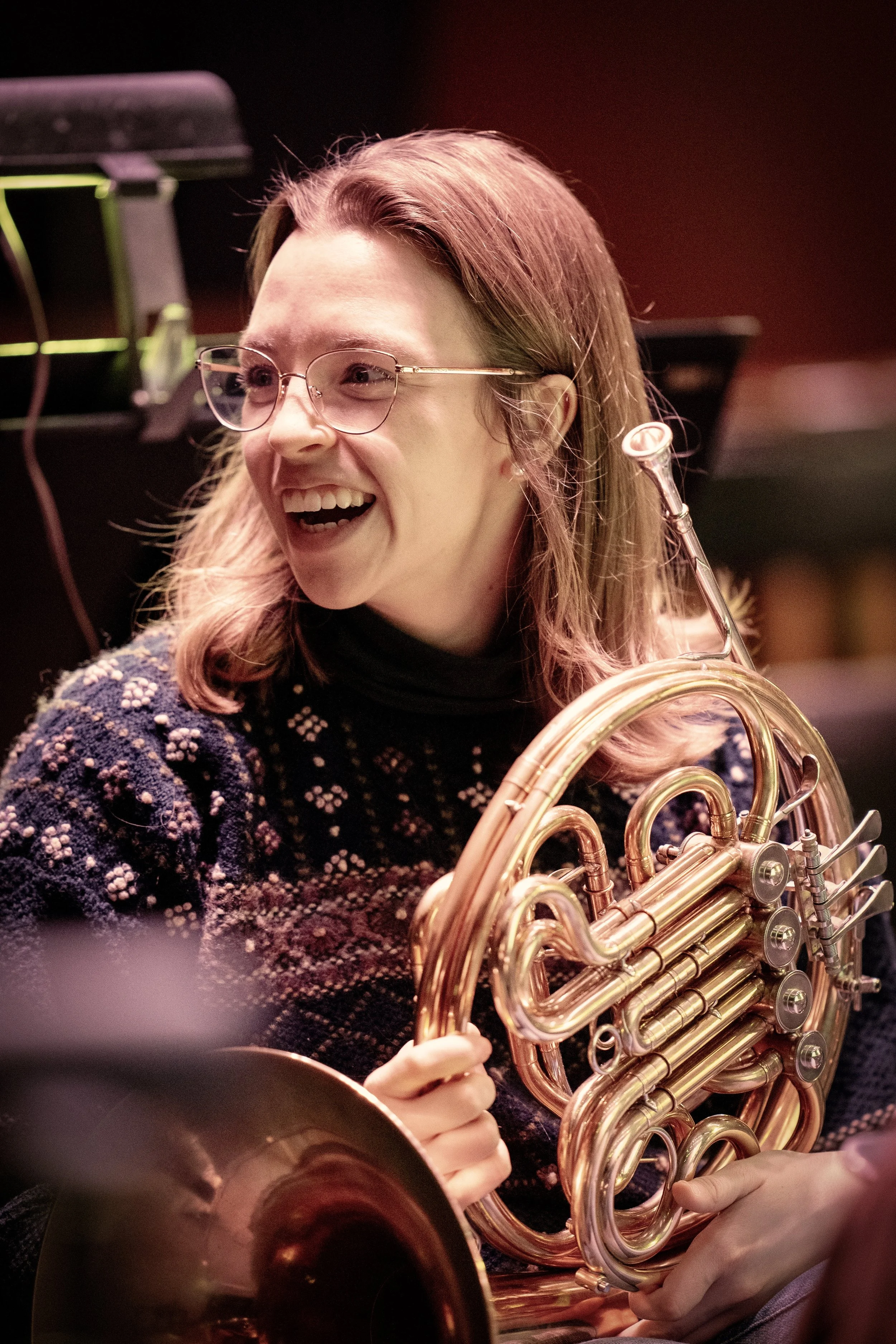 A woman with glasses and blonde hair holding a French horn, smiling and looking to her left in a music  concert hall during a rehearsal.