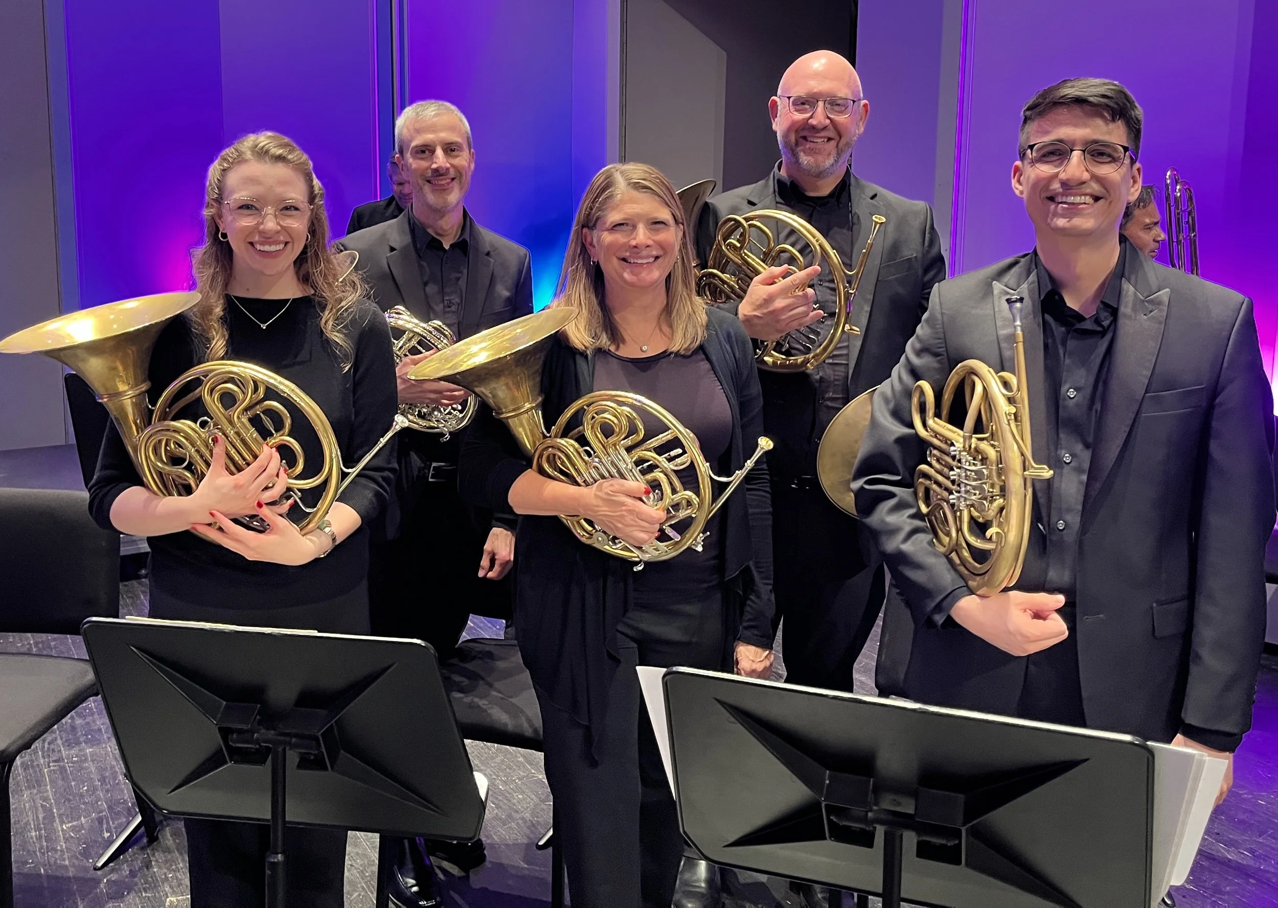 Group of six musicians holding French horns, standing in a concert hall with colorful lighting.