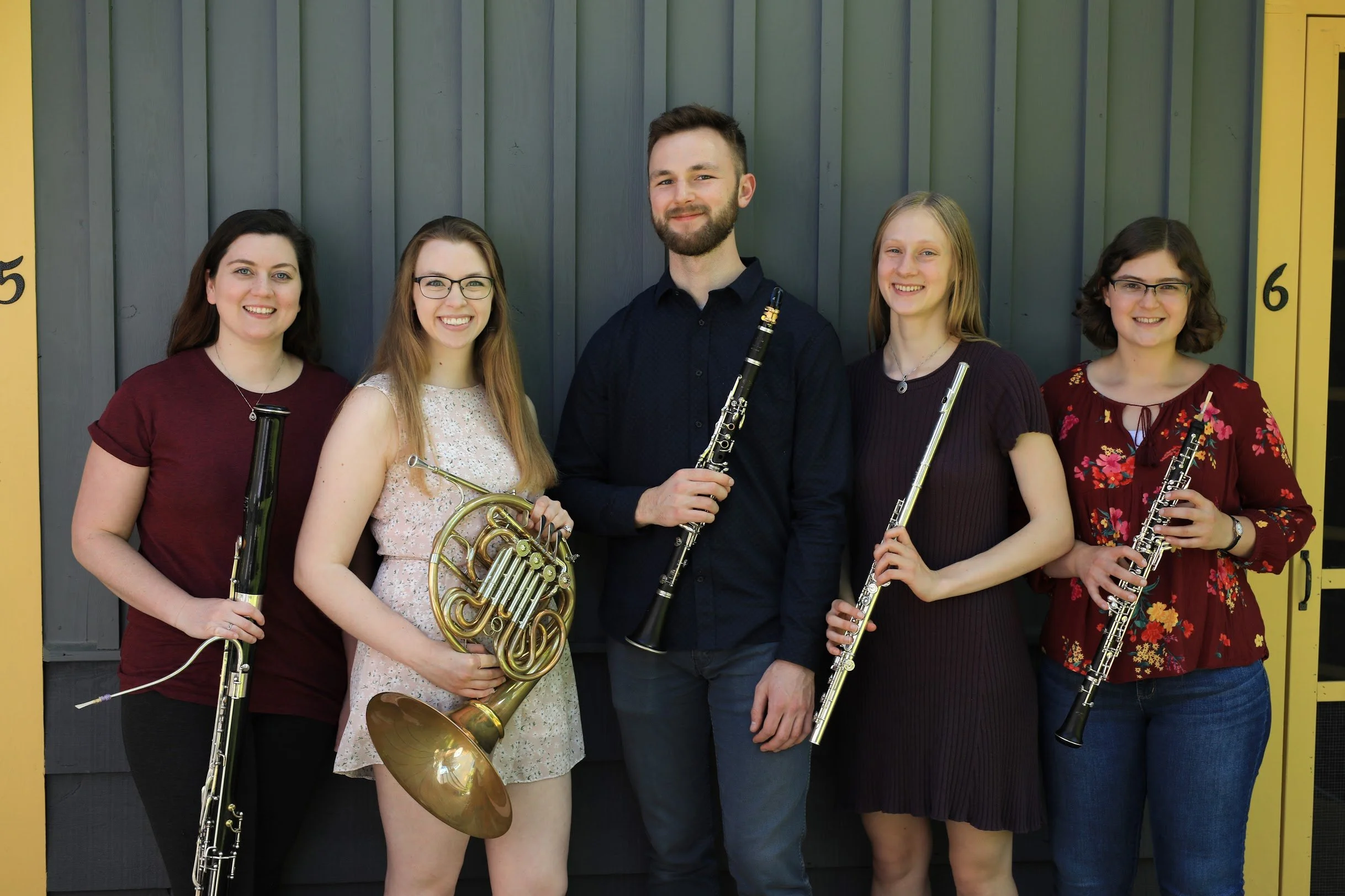 Group of five young women and one young man holding musical instruments bassoon, French horn, clarinet, flute, and oboe, standing in front of a gray wall with yellow accents.