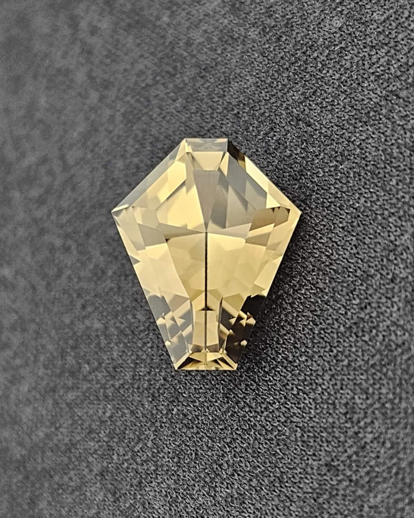 A clear, cut diamond-shaped crystal with faceted sides on a dark textured surface.