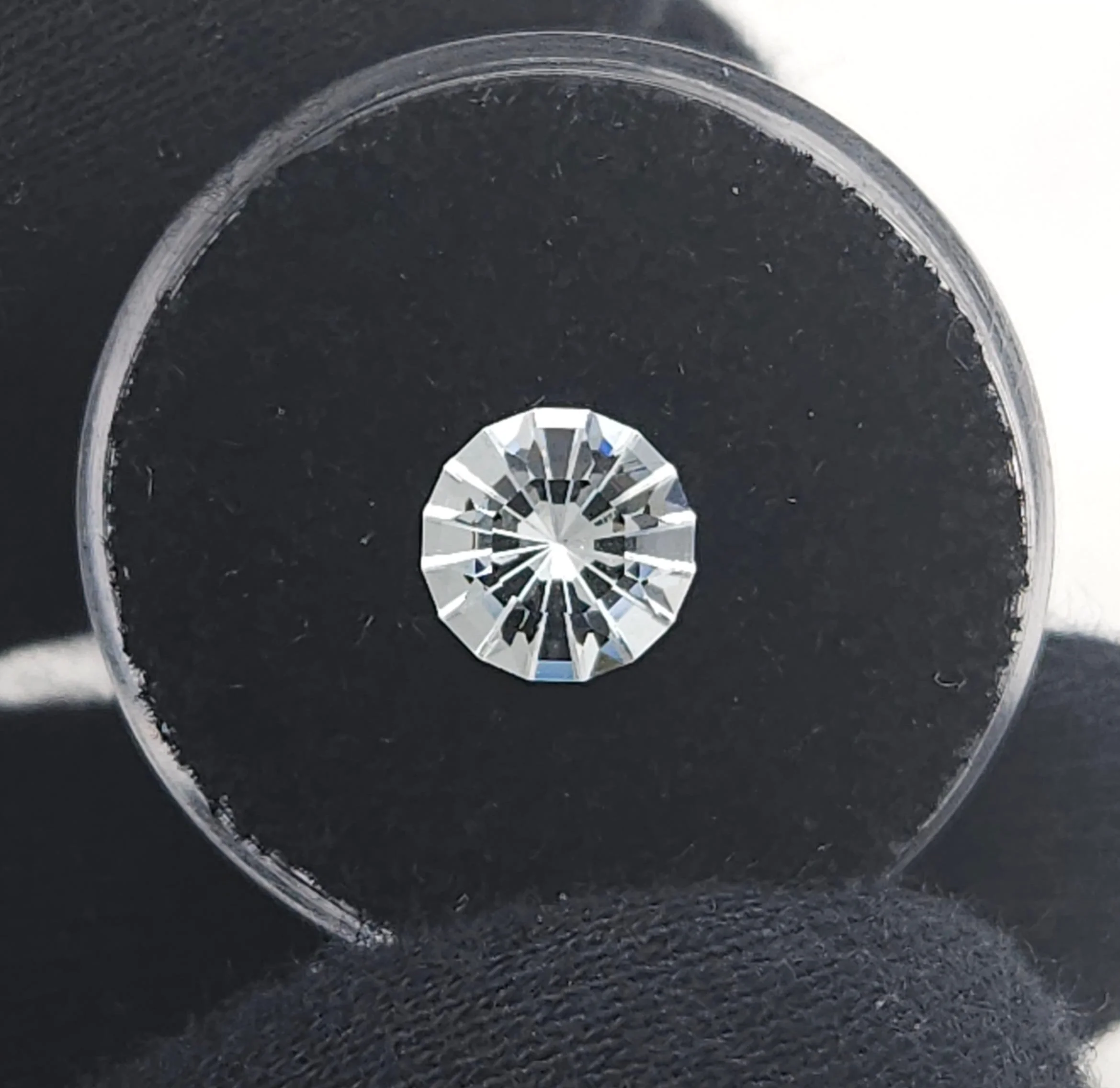 A round, brilliantly faceted clear glass gemstone with a detailed frosted starburst cut, displayed on a black velvet cushion inside a clear gem jar.