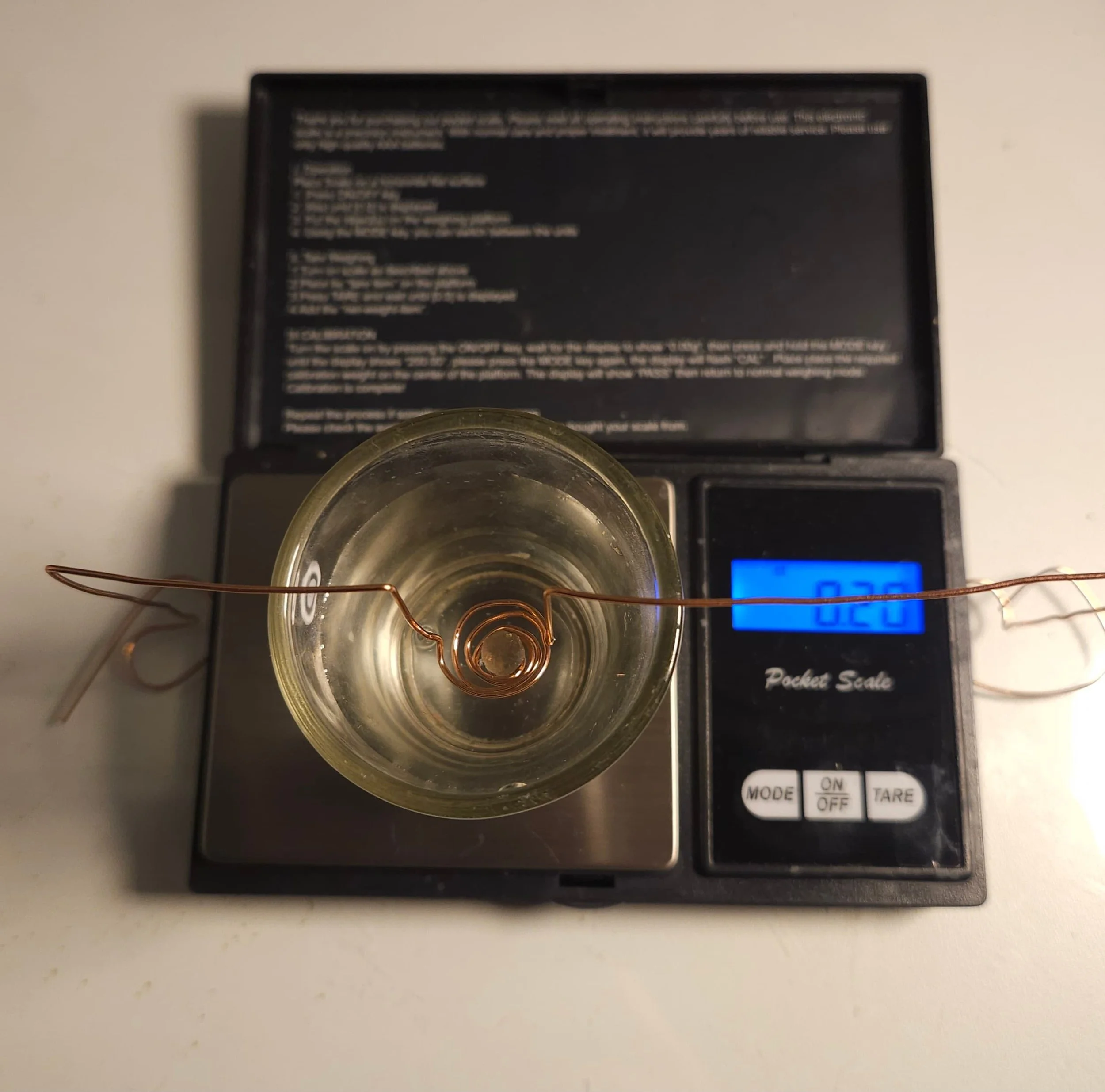 A digital pocket scale with a glass container on top, displaying a weight of 0.20 grams. The scale has buttons labeled MODE, ON/OFF, and TARE, and is placed on a white surface.
