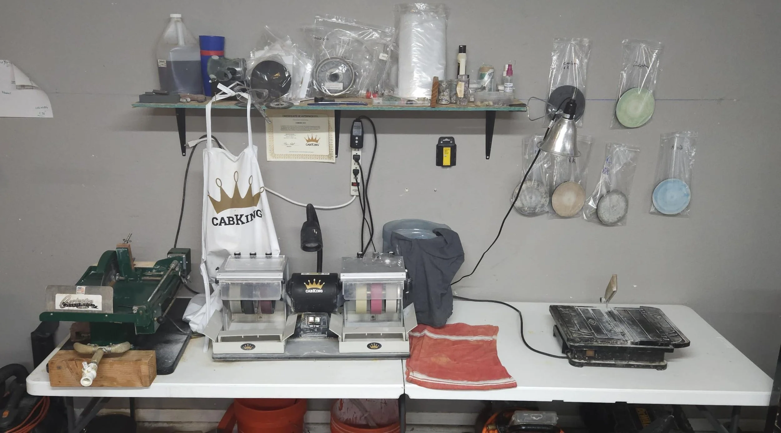 Workstation with baking equipment, including a dough sheeter, a heat sealer, a laminated towel, shelves with various tools, and decorative plates in plastic sleeves on the wall.