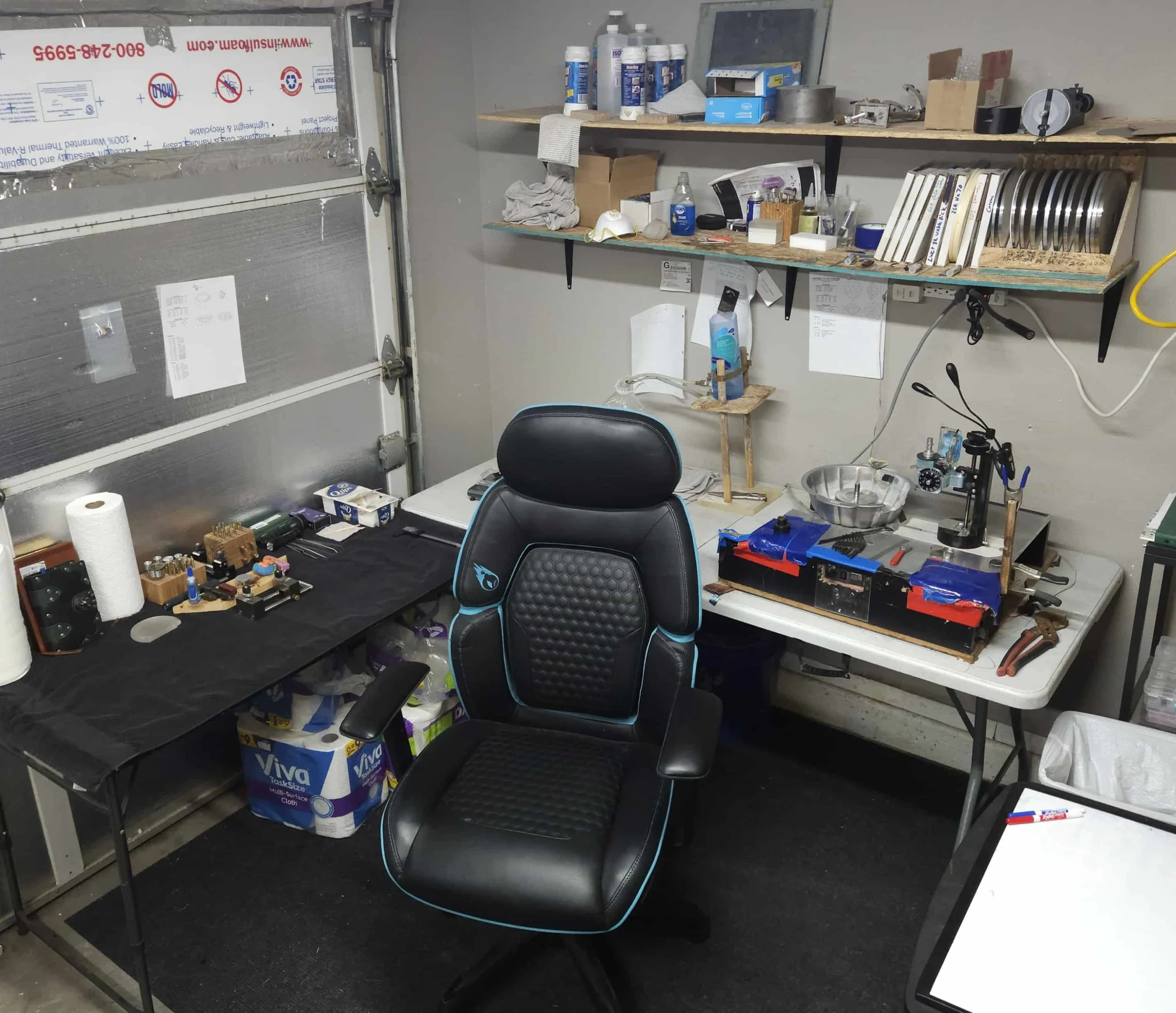 Workshop workspace with a black ergonomic chair, a workbench with tools and equipment, shelves with various supplies, and a garage door.