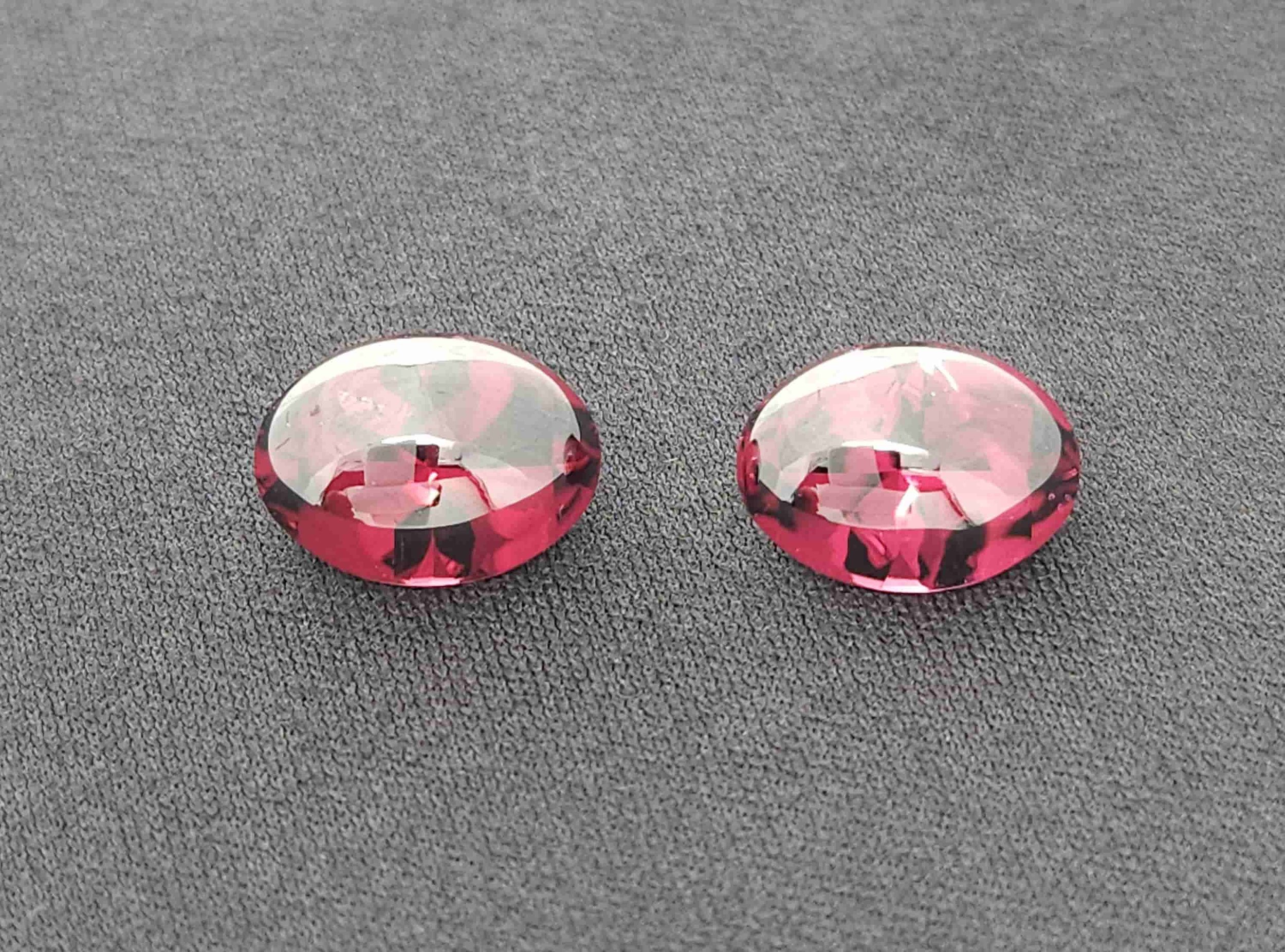 Two faceted and cabbed, oval-shaped rhodolite garnet gemstones showcasing a vibrant pinkish-red hue side by side. The matching pair rests on a textured, dark gray fabric surface.