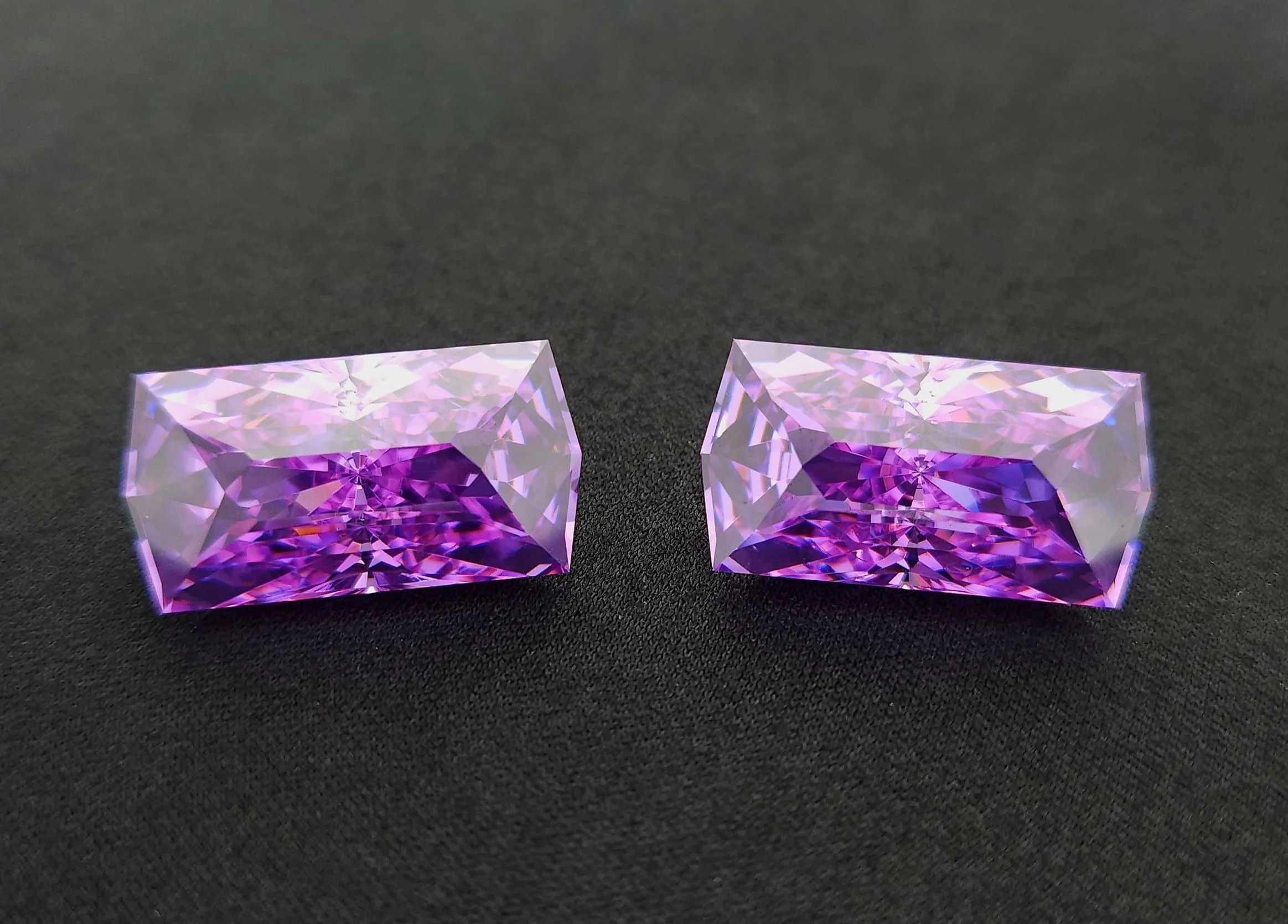 Two square-cut, purple and pink gemstones with intricate facets, placed on a black surface.