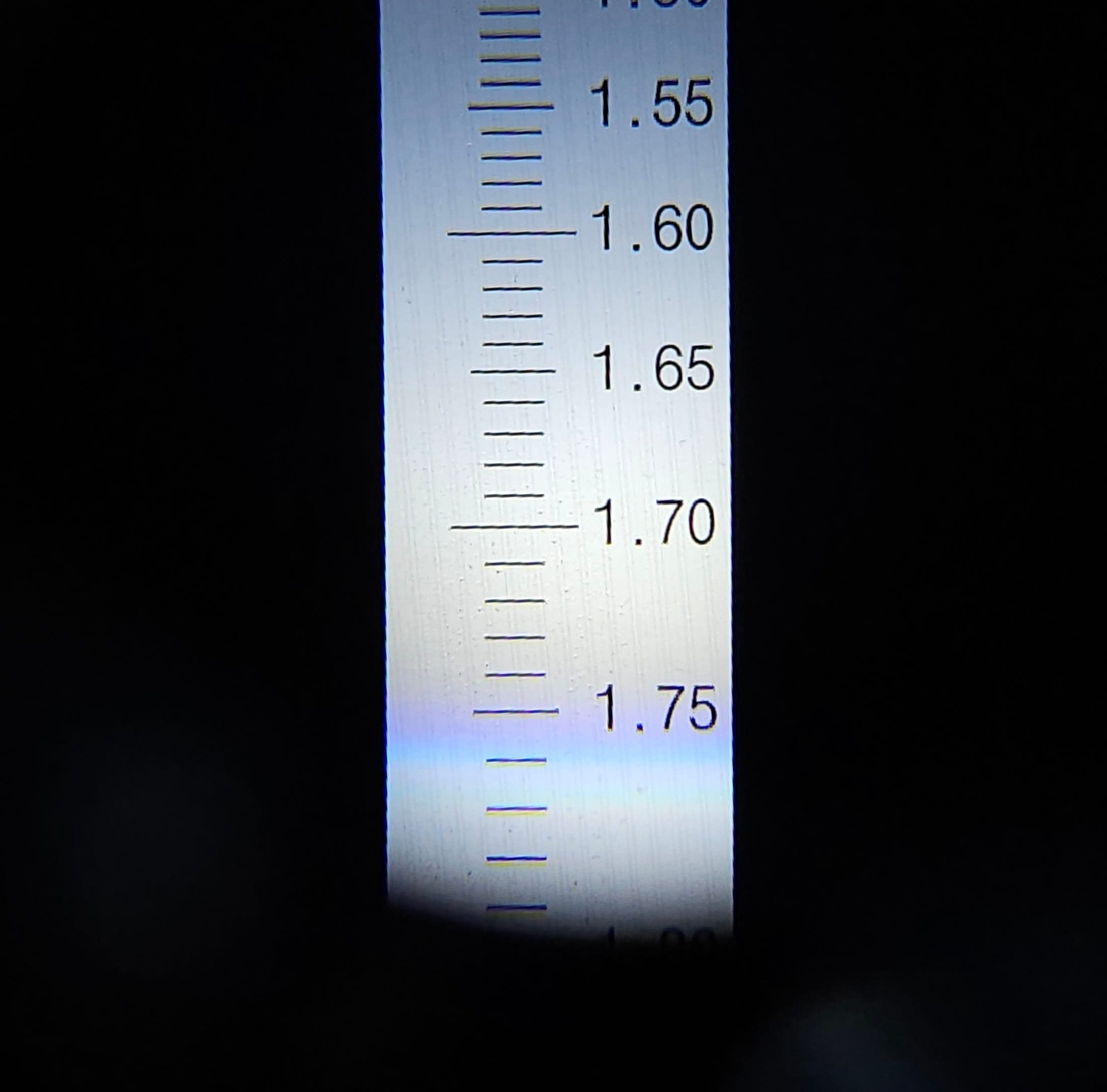 Close-up of a refractometer reading with black and yellow markings, showing measurements from 1.55 to 1.75. The background is black.