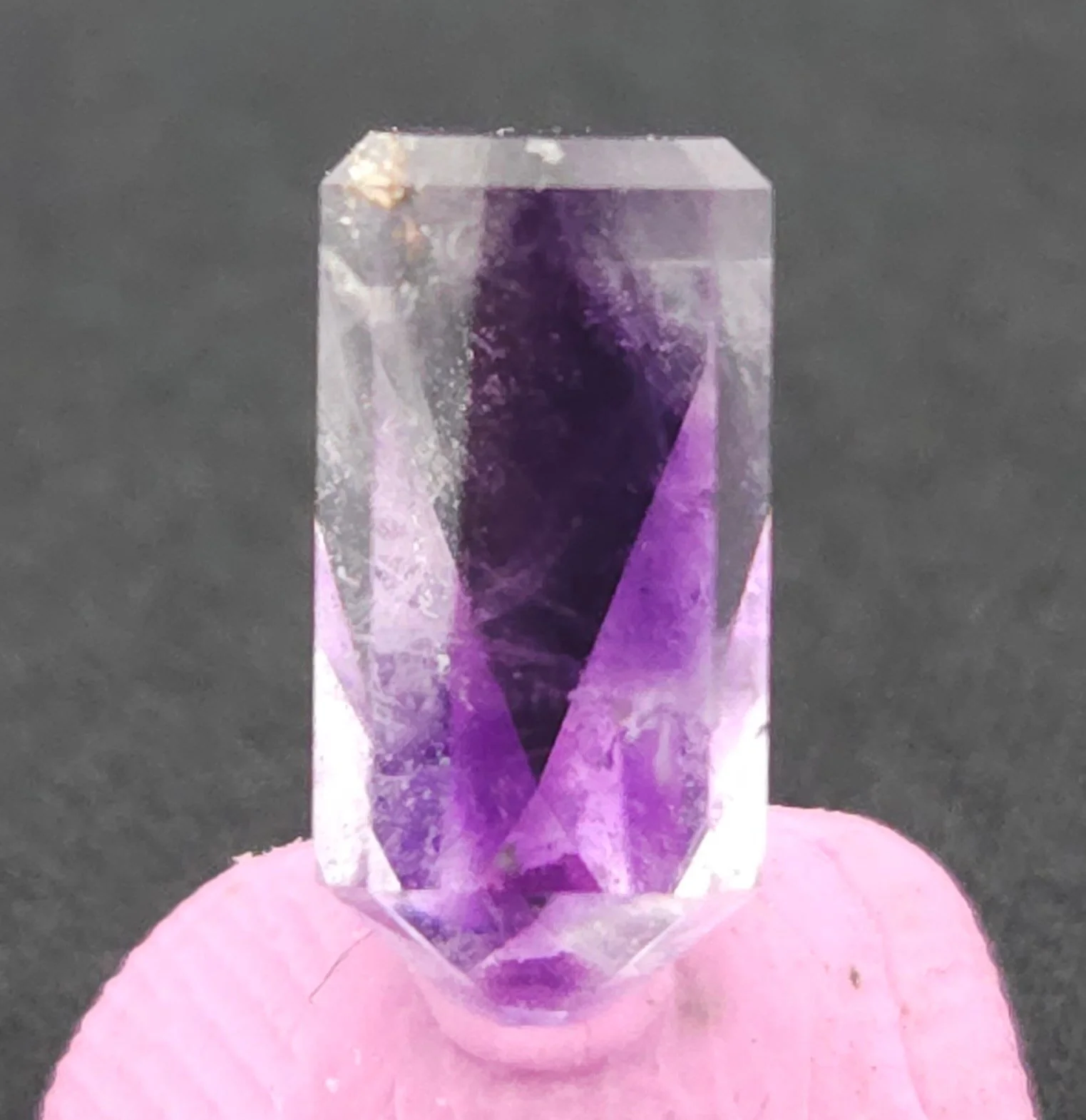 A faceted, rectangular Four Peaks amethyst gemstone with a sharp, dark purple V-shaped phantom pattern. The crystal is mounted on a pink base against a dark gray background.