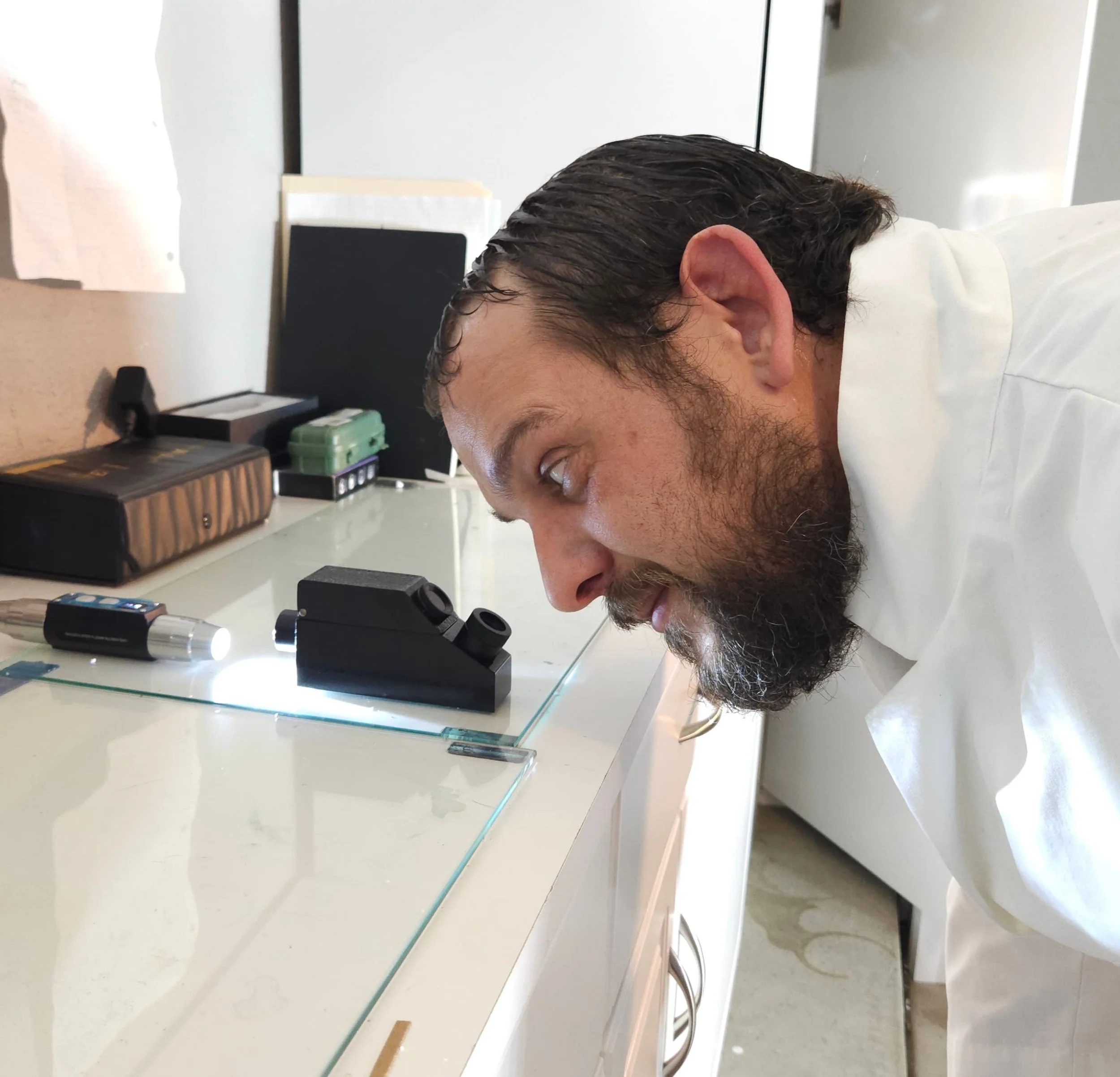 Ian Perry from The Lapidary Office looking into a refractometer in a lab environment, with various office supplies and equipment visible in the background.