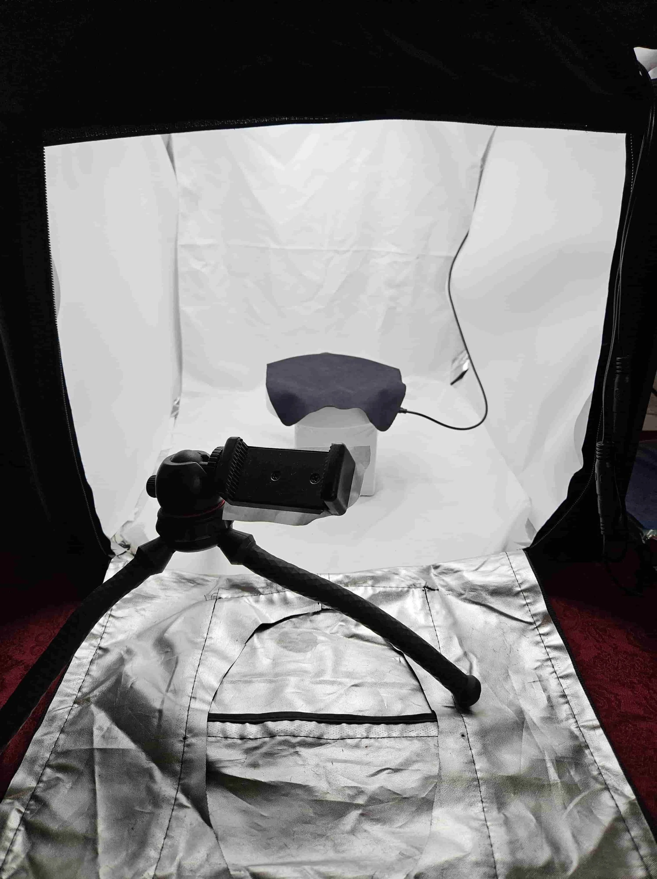 A small photography studio setup with a reflective backdrop and a camera on a tripod, focusing on a dark cloth-covered object on a white pedestal, connected to a power source.