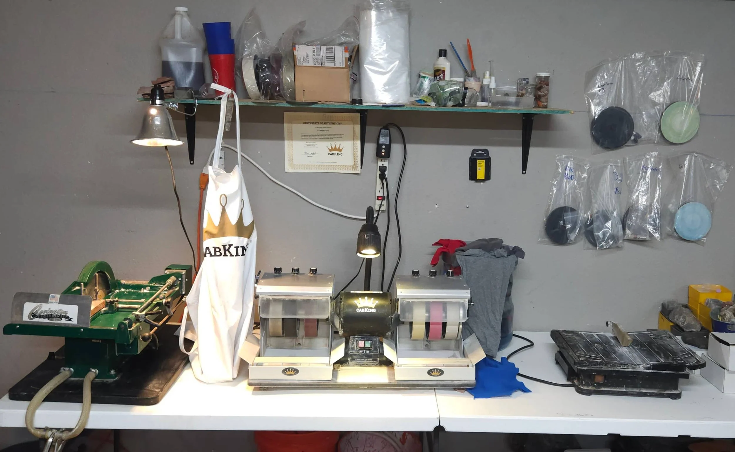 A workshop workbench featuring a six inch cabochon machine, a ten inch slab saw, and a 4 inch trim saw with lapidary accessories on a shelf and pinned on a wall.