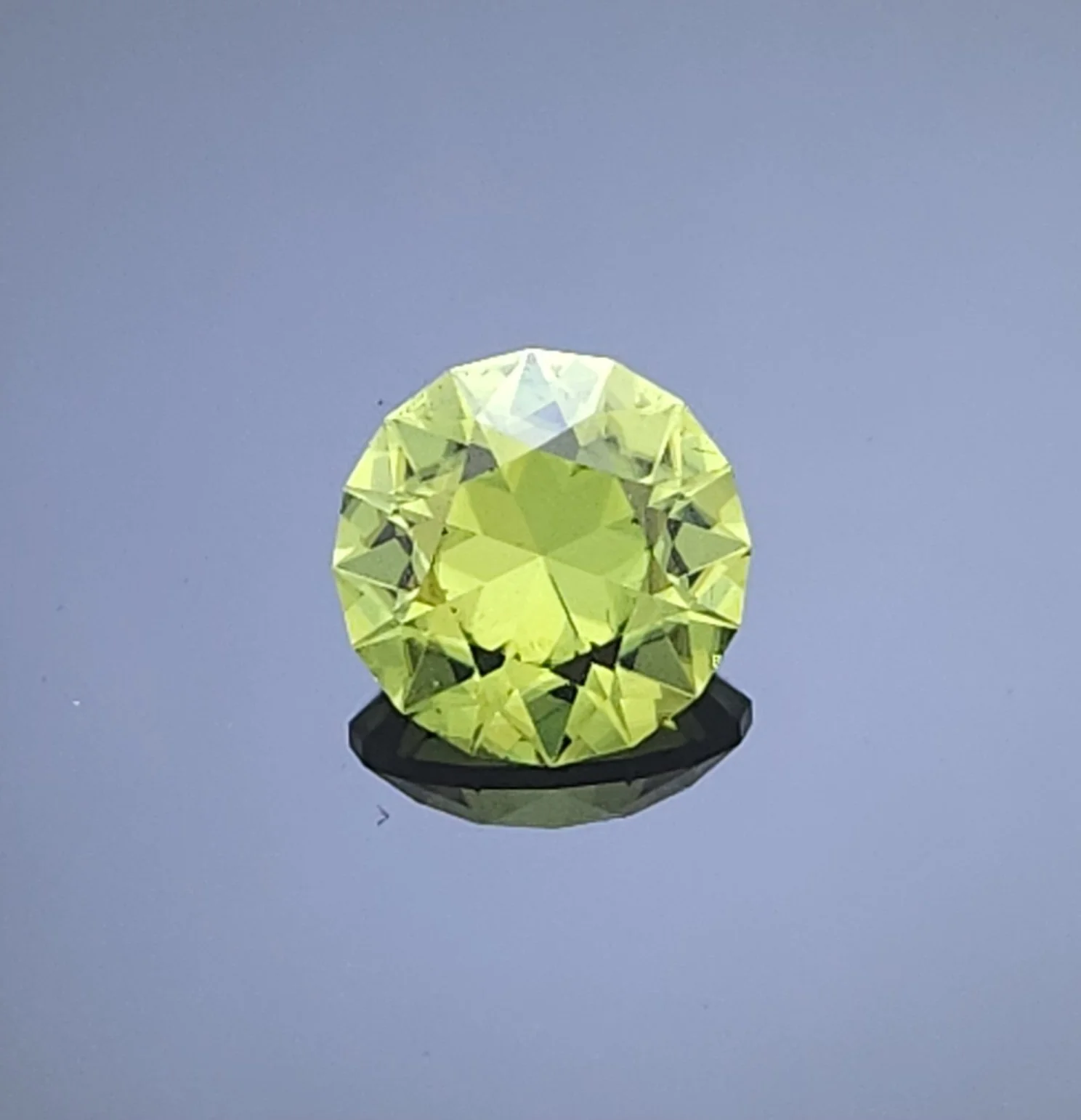 Standard round briliant cut Peridot resting on a grey reflective surface.