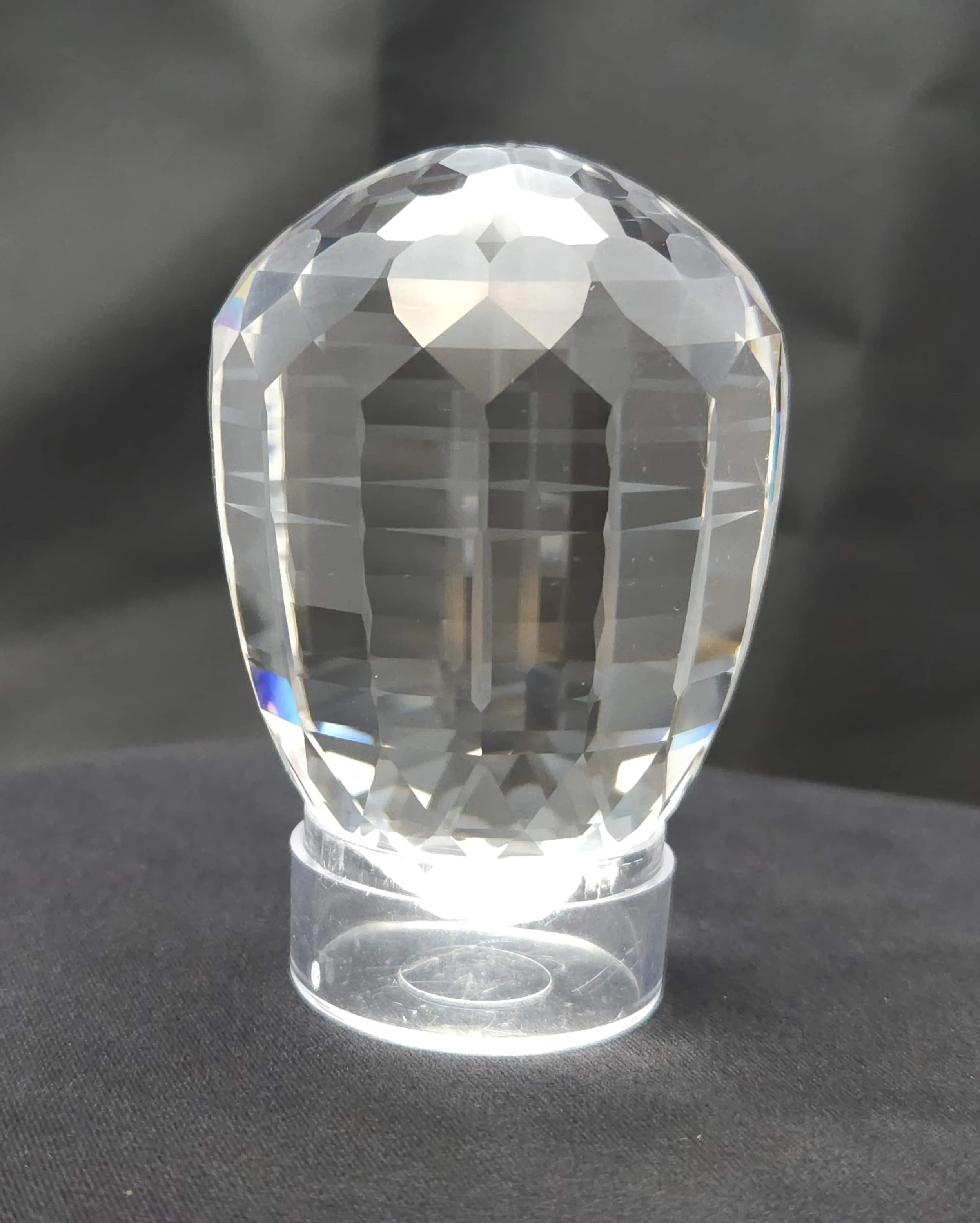 A large, precision-faceted Brazilian quartz crystal egg showing brilliant geometric reflections. The clear gemstone stands upright on a small plastic display ring against a black background.