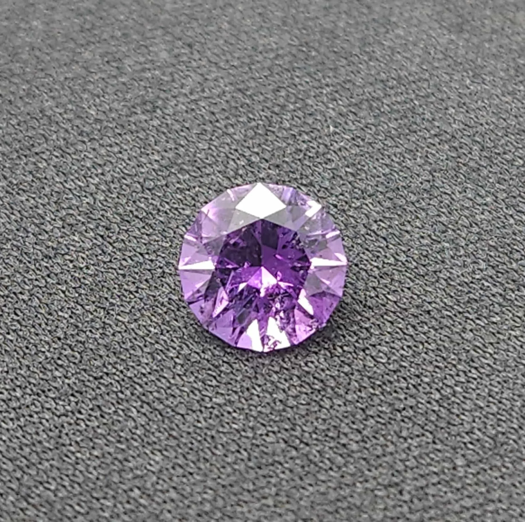 A brilliant, round-cut Four Peaks amethyst gemstone displaying bright violet facets and deep purple reflections. The circular gem rests centered on a textured, dark gray fabric background.